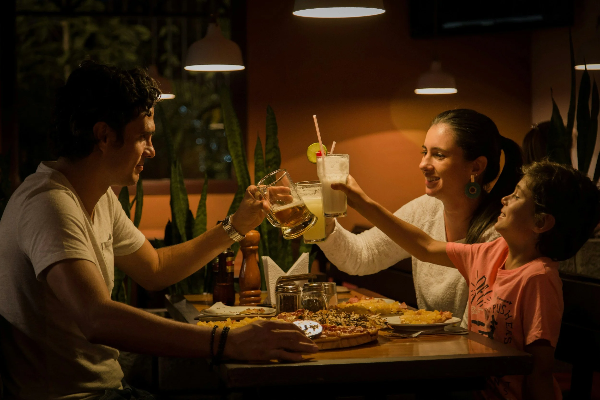 Family toasting with drinks at a restaurant table with pizza and other dishes.