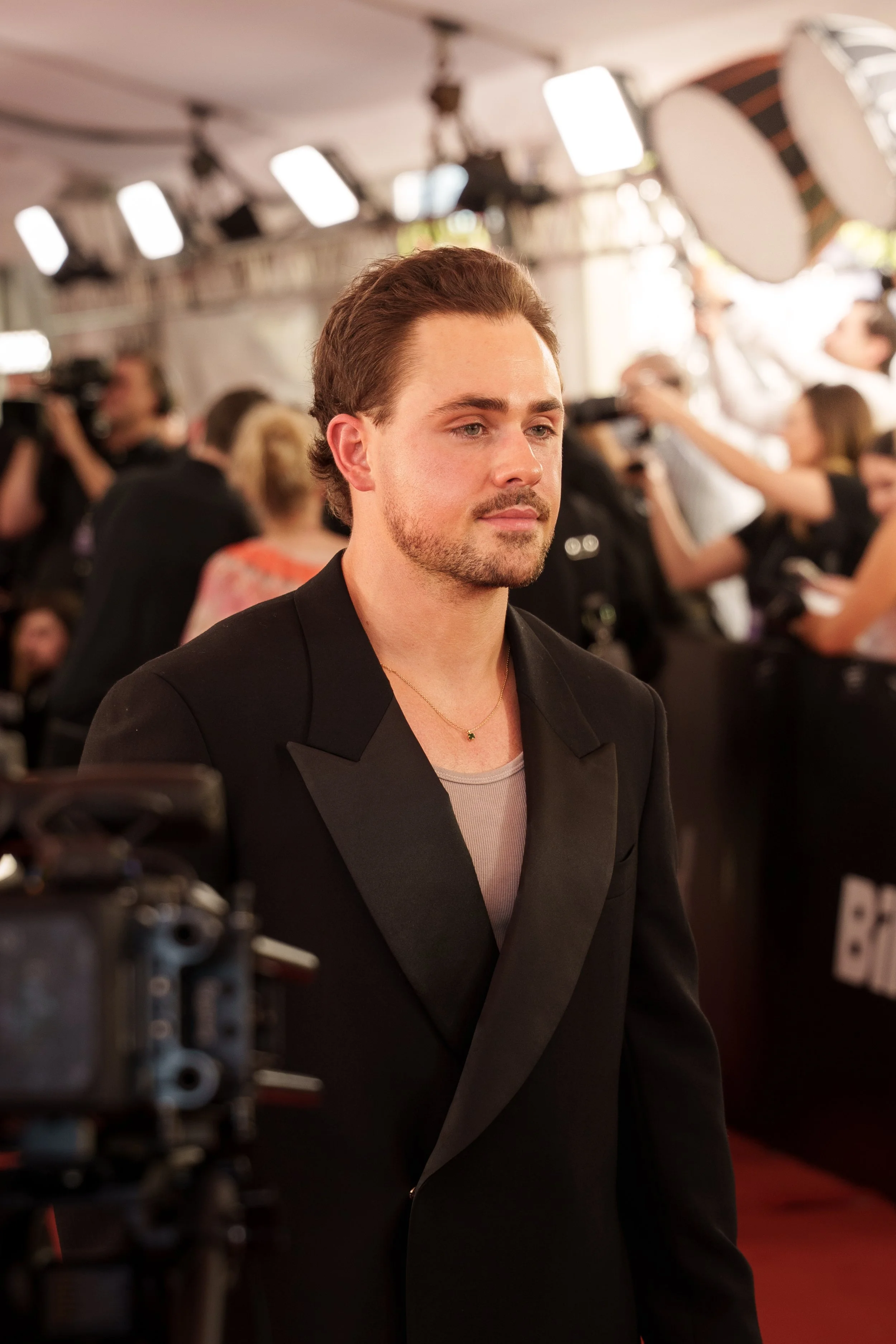 A man with light skin and brown hair styled back, wearing a black blazer over a light gray shirt, standing on a red carpet at an event, surrounded by photographers and people in the background.