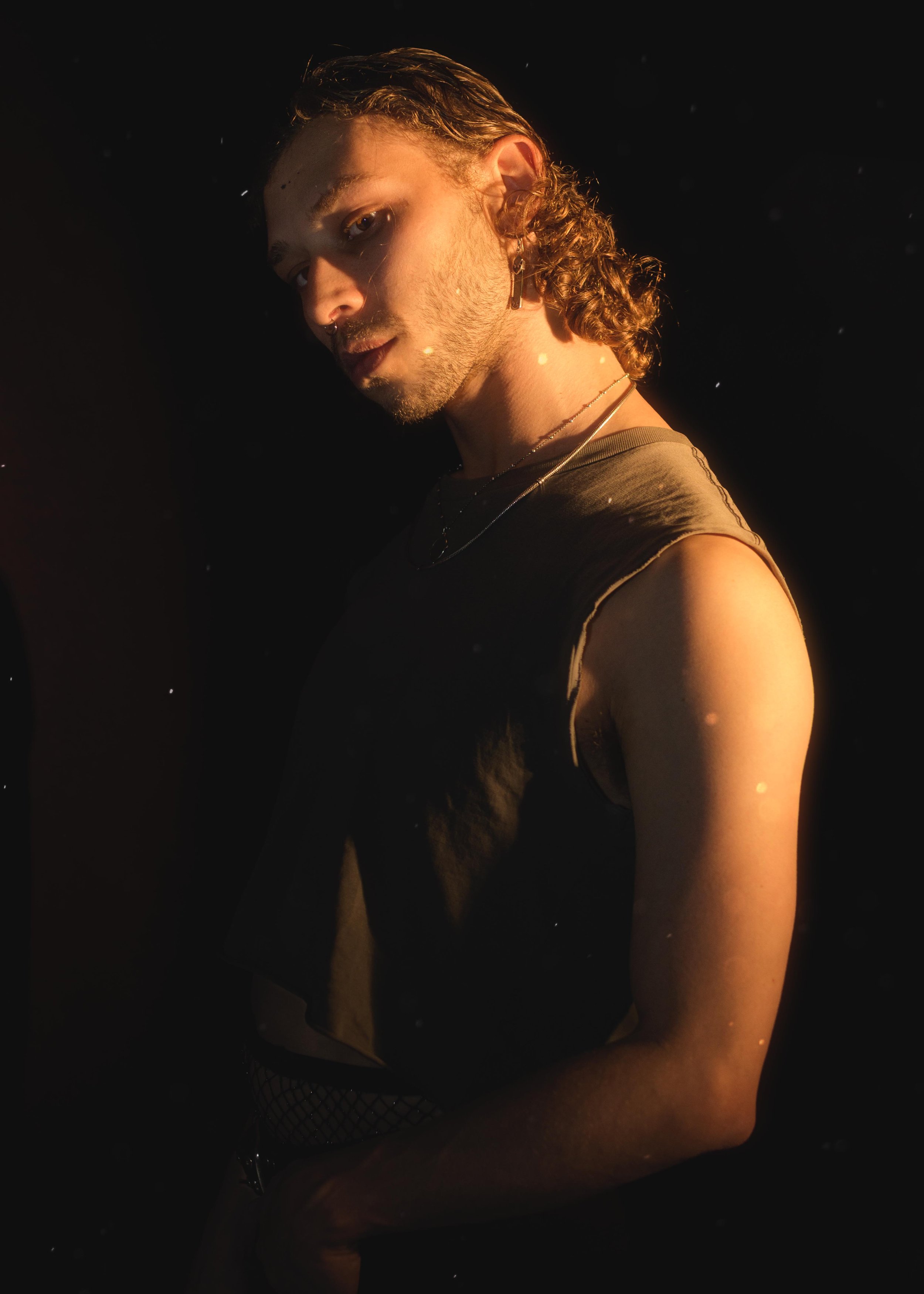 A young man with wavy hair and earrings is wearing a black sleeveless shirt and fishnet pants, posing against a dark background with warm lighting highlighting his profile.