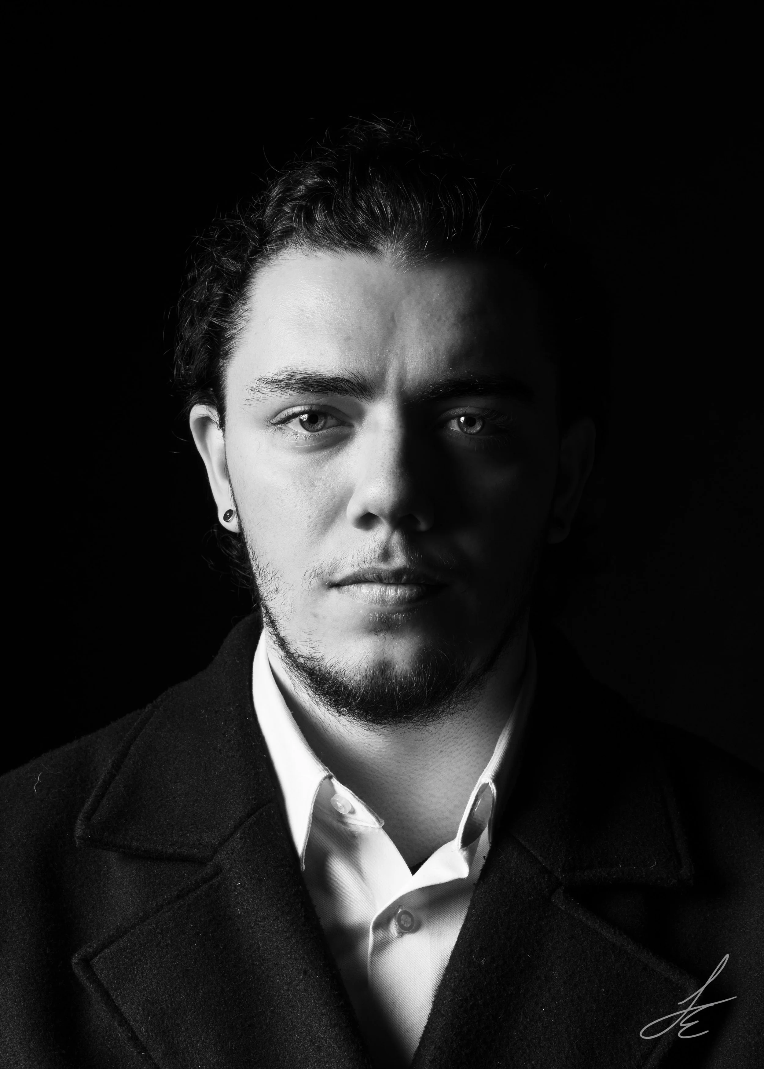 Black and white portrait of a young man with curly hair, facial hair, wearing a coat and dress shirt, looking directly at the camera with a serious expression, against a dark background.