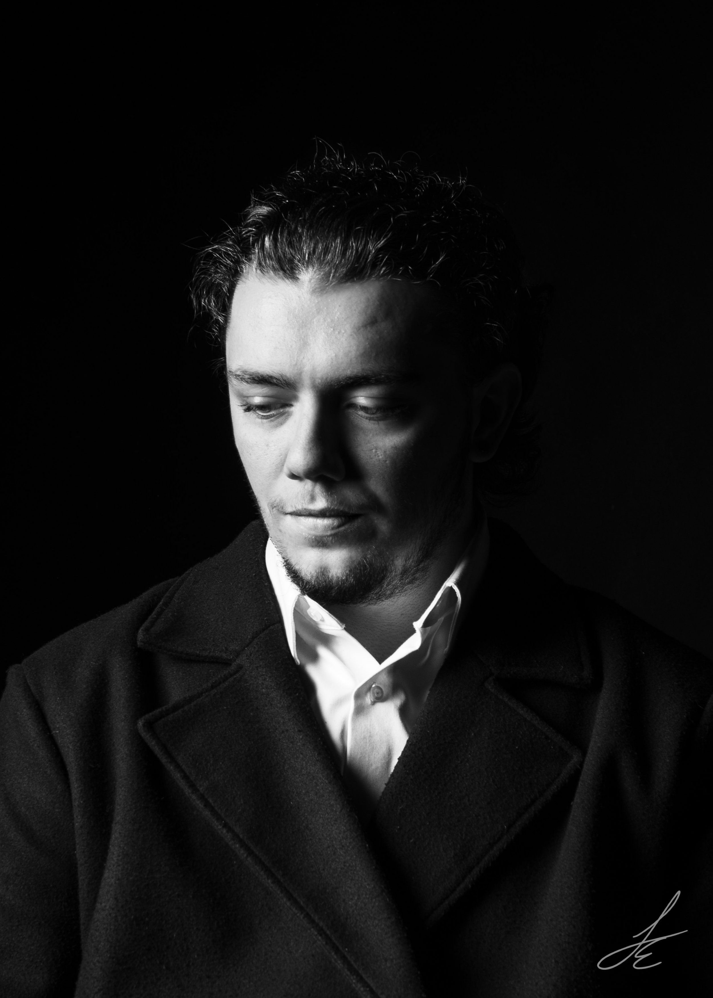 Black and white portrait of a young man with his eyes closed, wearing a dark coat over a white shirt, against a dark background.