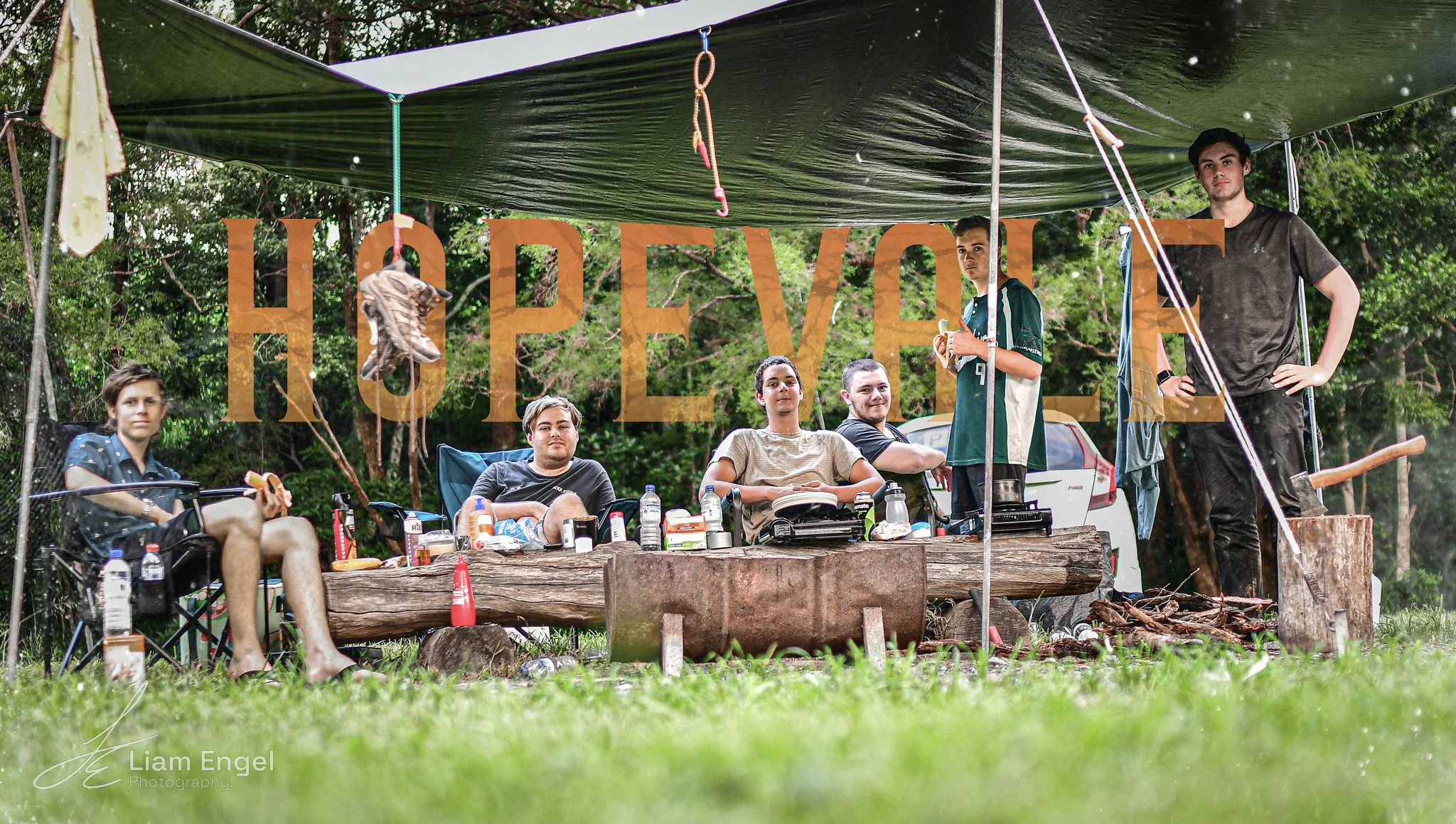 Group of young people gathering outdoors under a canopy, sitting and standing around with picnic supplies, with a wooden table and nature background. The word 'HOPE' is written across the image.