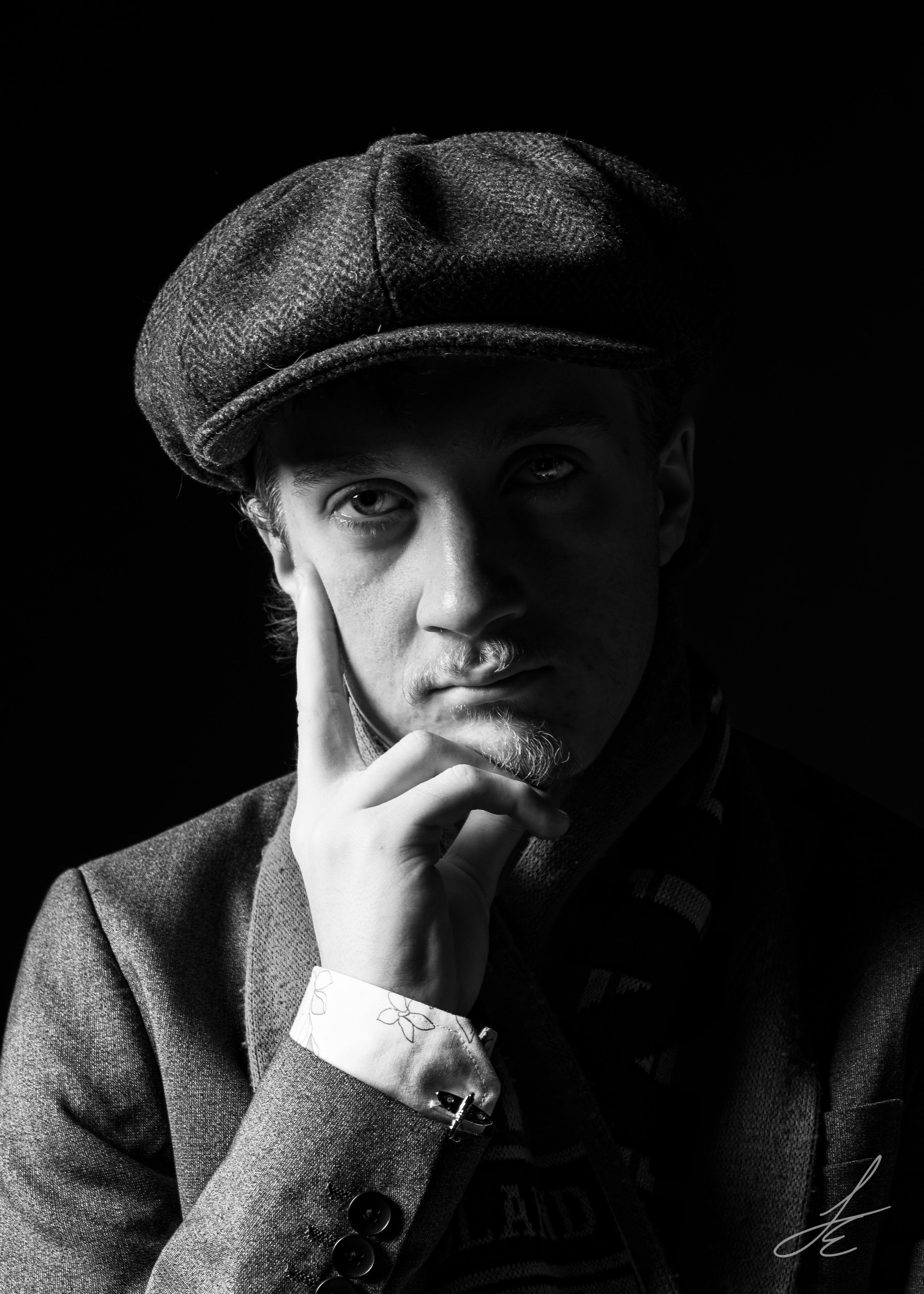 A black and white portrait of a young man wearing a flat cap, with a thoughtful expression, resting his face on his hand, dressed in a blazer and patterned shirt, against a dark background.
