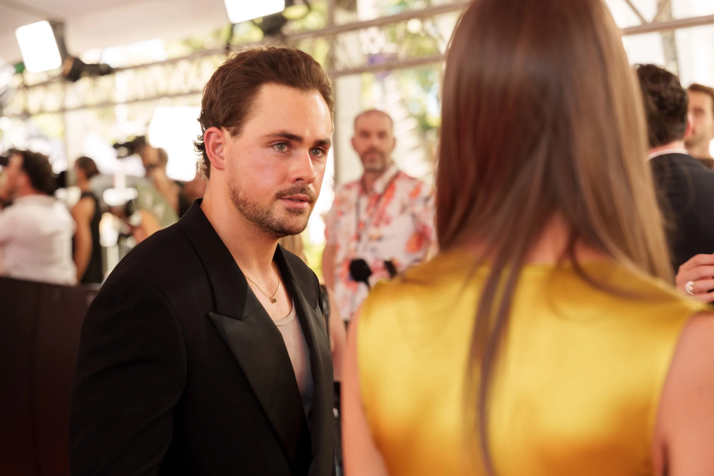 A man with brown hair and a beard, dressed in a black blazer, is looking at a woman in a yellow top during a social gathering or event. Other people are visible in the background.