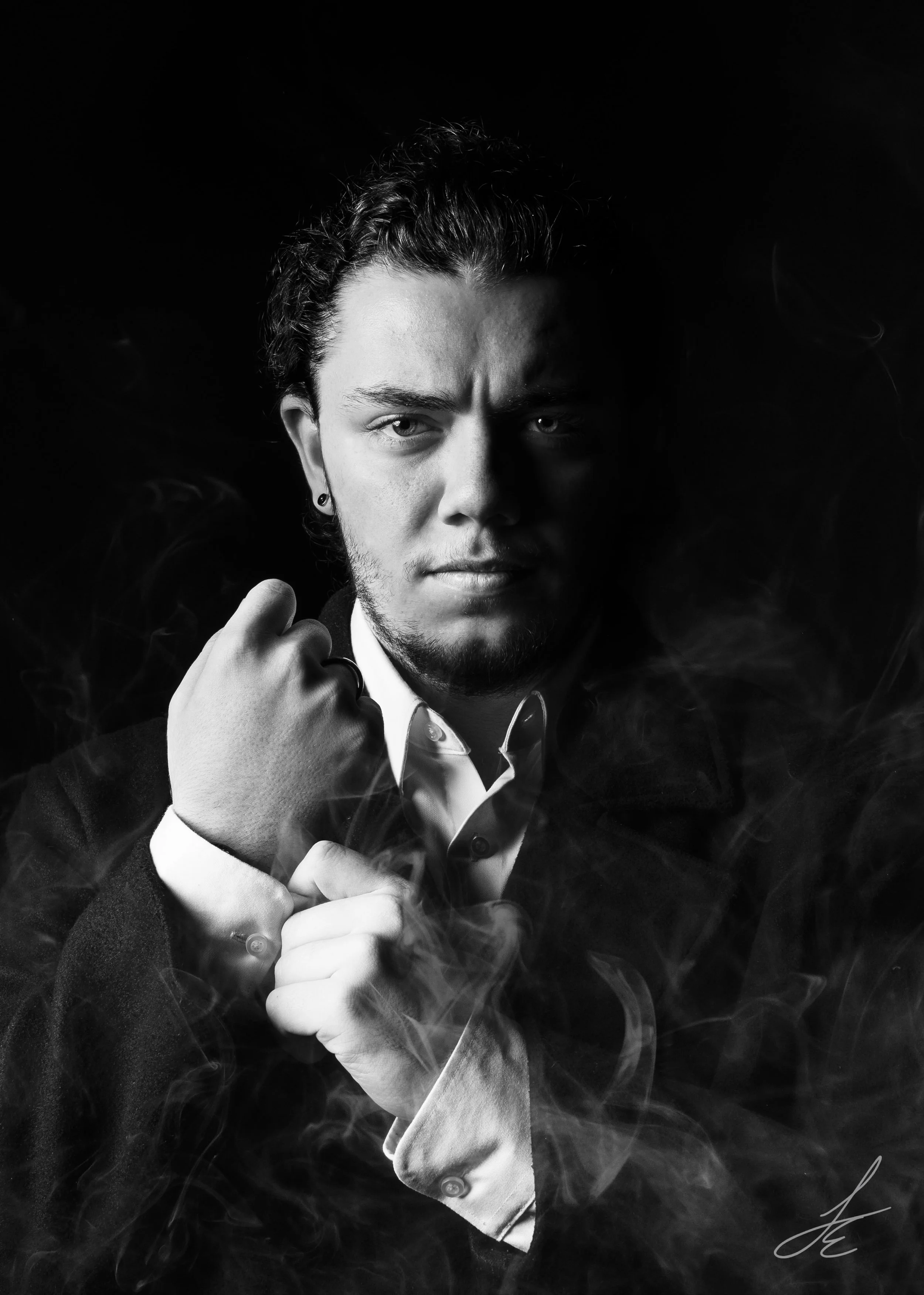 Black and white portrait of a man with dark, curly hair, pointing his fist towards the camera, wearing a collared shirt and a dark vest, with smoke swirling around him, dark background.