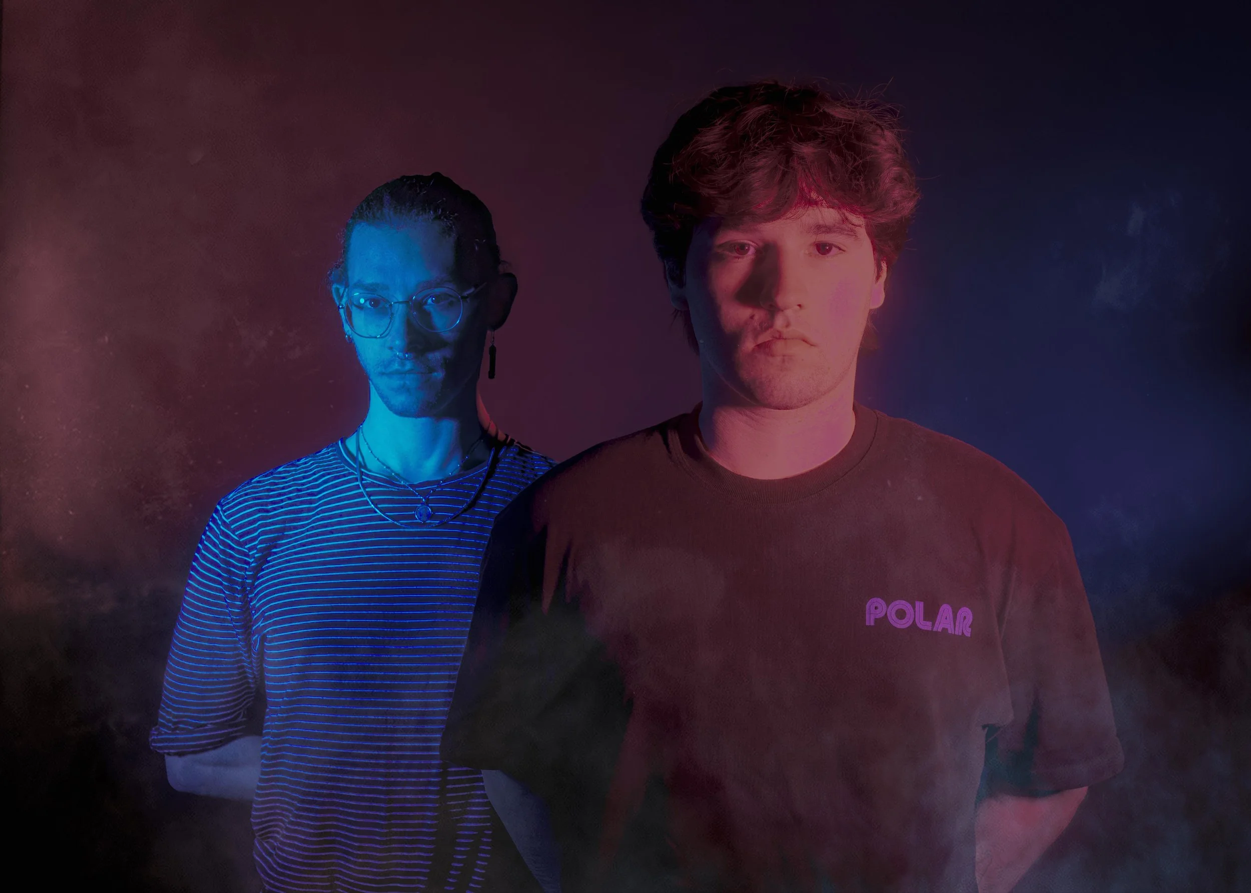 A man and a woman are standing together with colorful lighting and fog. The man is wearing a black shirt with purple text that reads 'POLAR.' The woman is wearing glasses, earrings, necklaces, and a striped shirt.