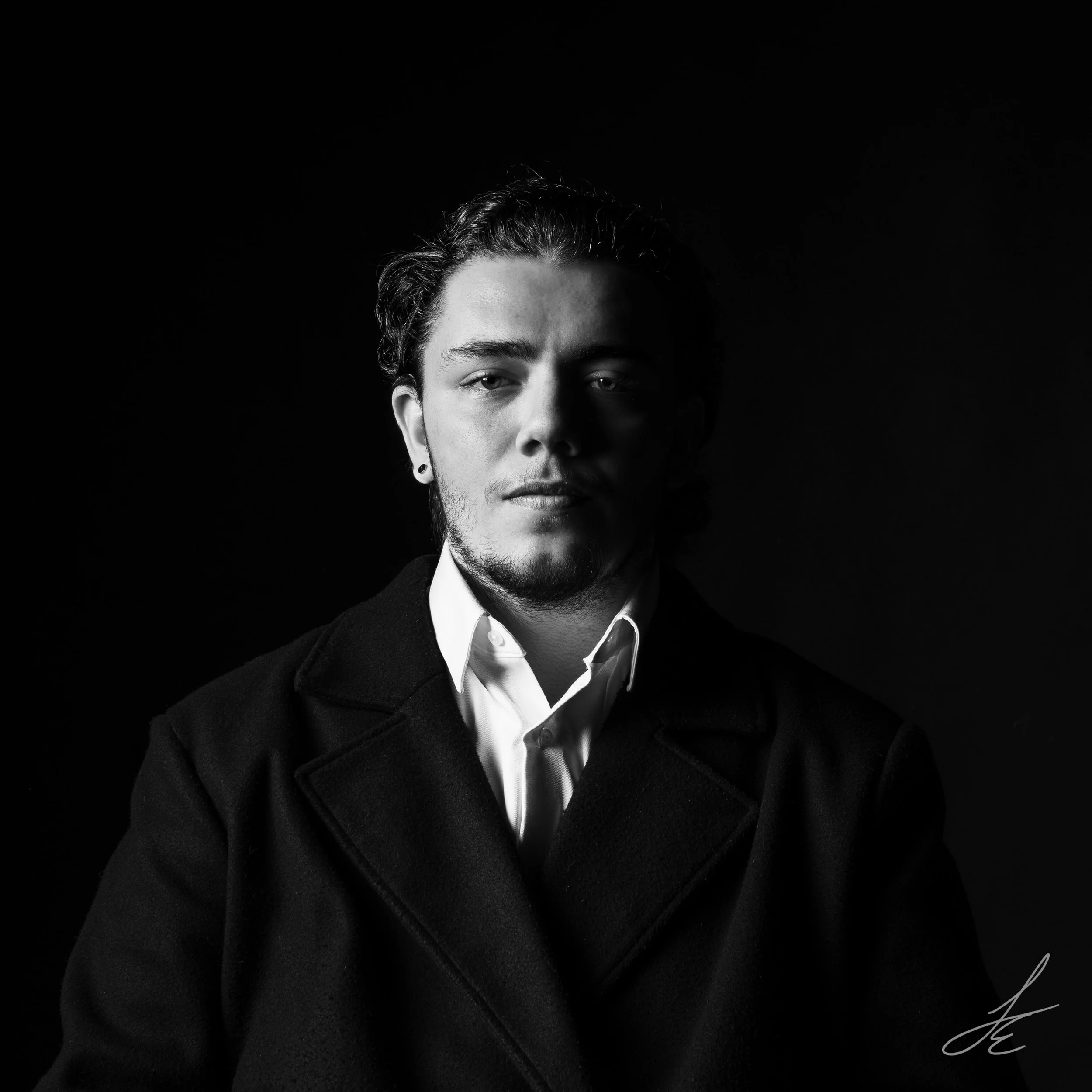 Black and white portrait of a young man with wavy hair, wearing a dark coat and a white shirt, against a dark background.