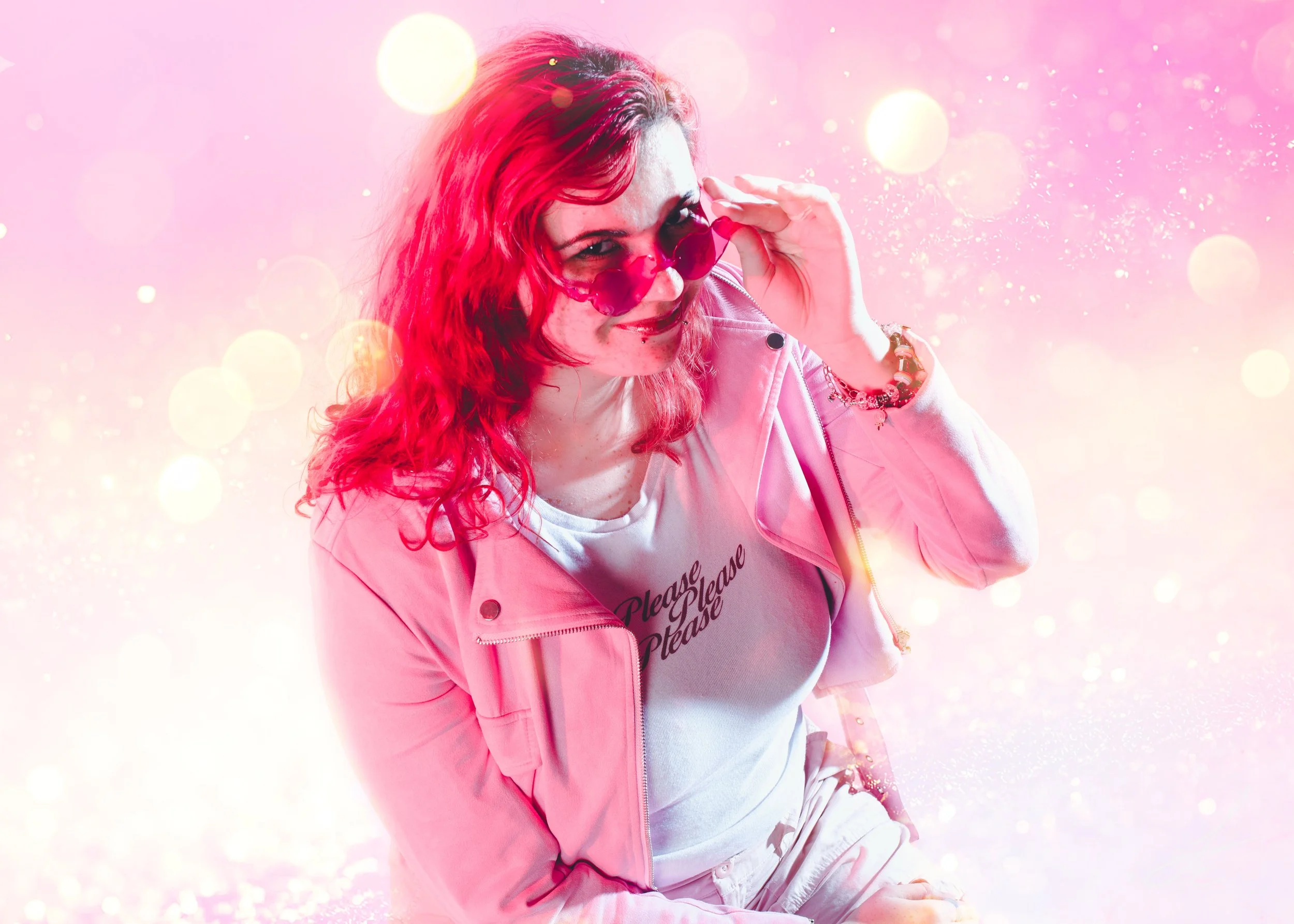 A woman with red hair wearing pink sunglasses, a pink jacket, and a white T-shirt with the text 'Please Steal' against a pink and pastel-colored sparkly background.