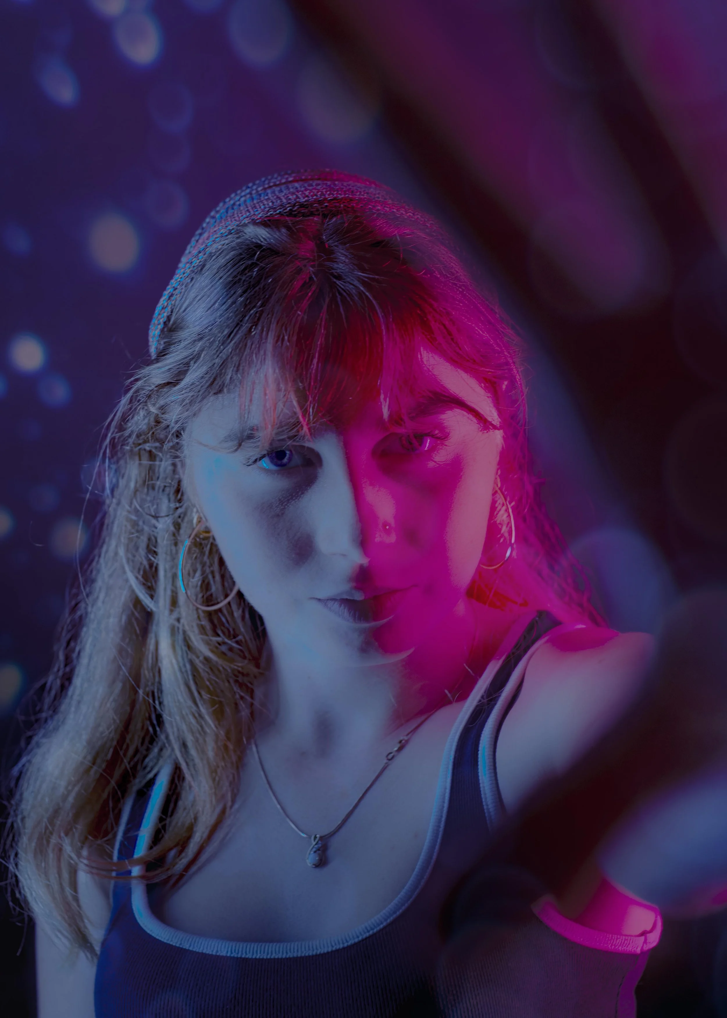 A young woman with blonde hair and one eye blue, wearing hoop earrings, a necklace, and a tank top, is taking a selfie with neon pink and blue lighting creating a colorful, vibrant background.