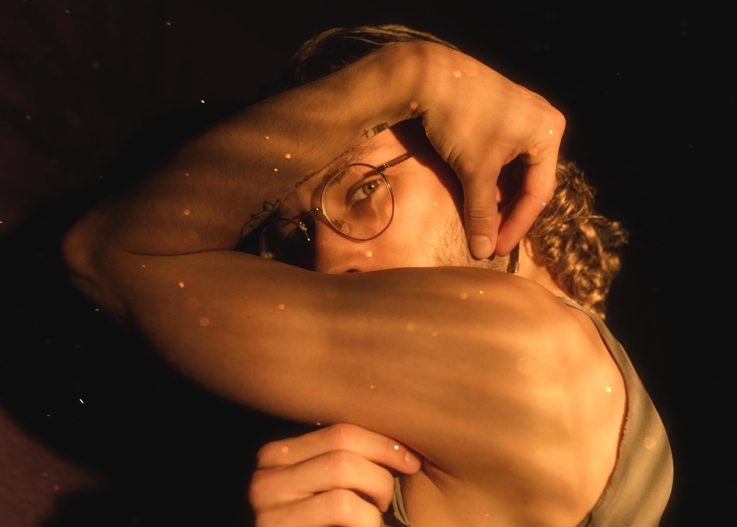 Close-up of a young man with glasses, resting his forehead on his arm while lying down in a dark setting with golden specks of light.