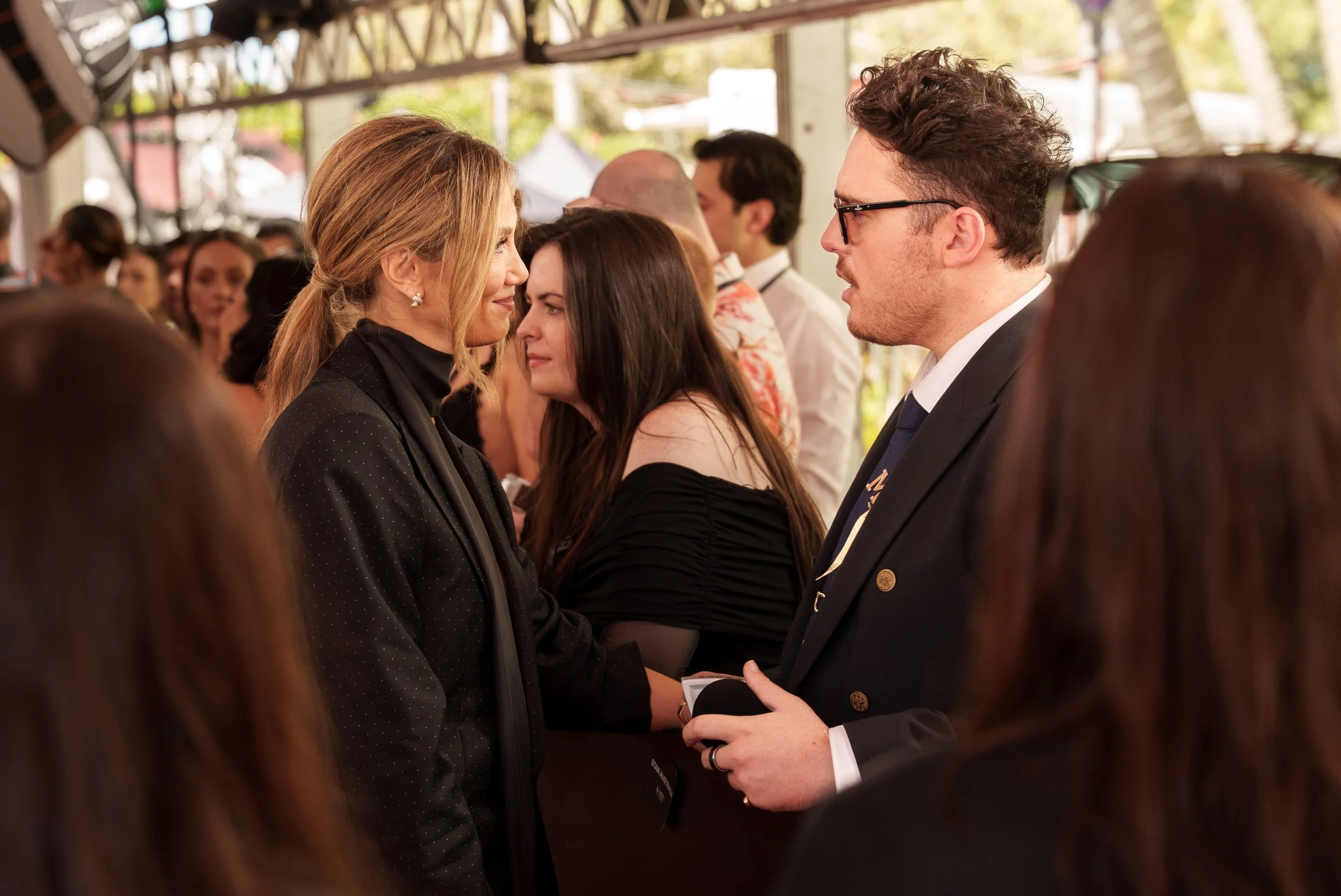 A group of people at an outdoor event, with three people in focus engaging in conversation: a woman in a black jacket, a woman with dark hair in a black off-the-shoulder top, and a man in glasses and a suit.