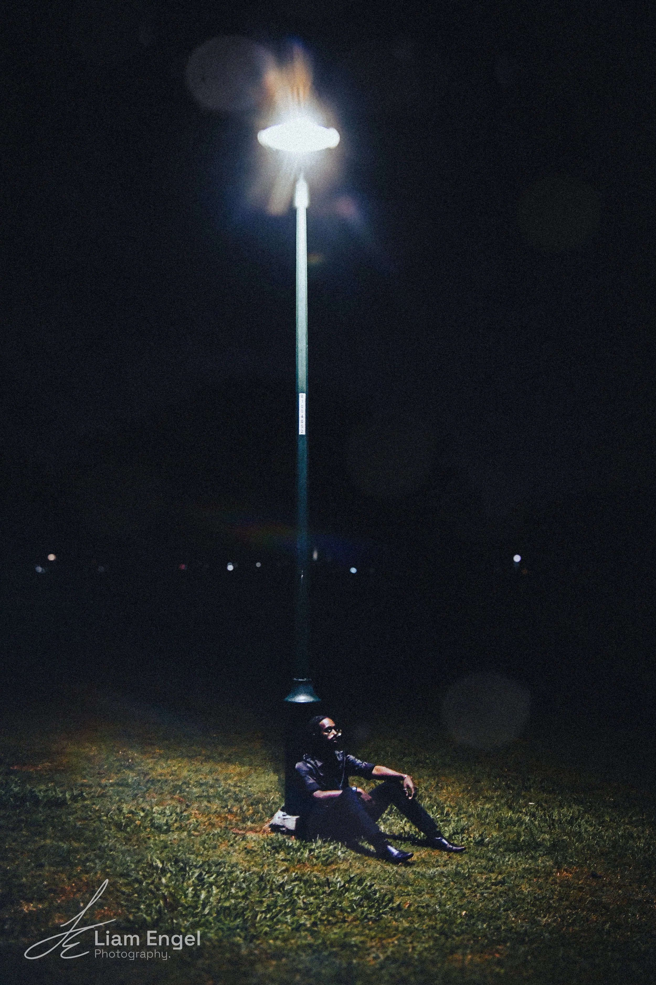A person is sitting on the grass at night, under a tall streetlamp that is turned on, illuminating them against the dark background.