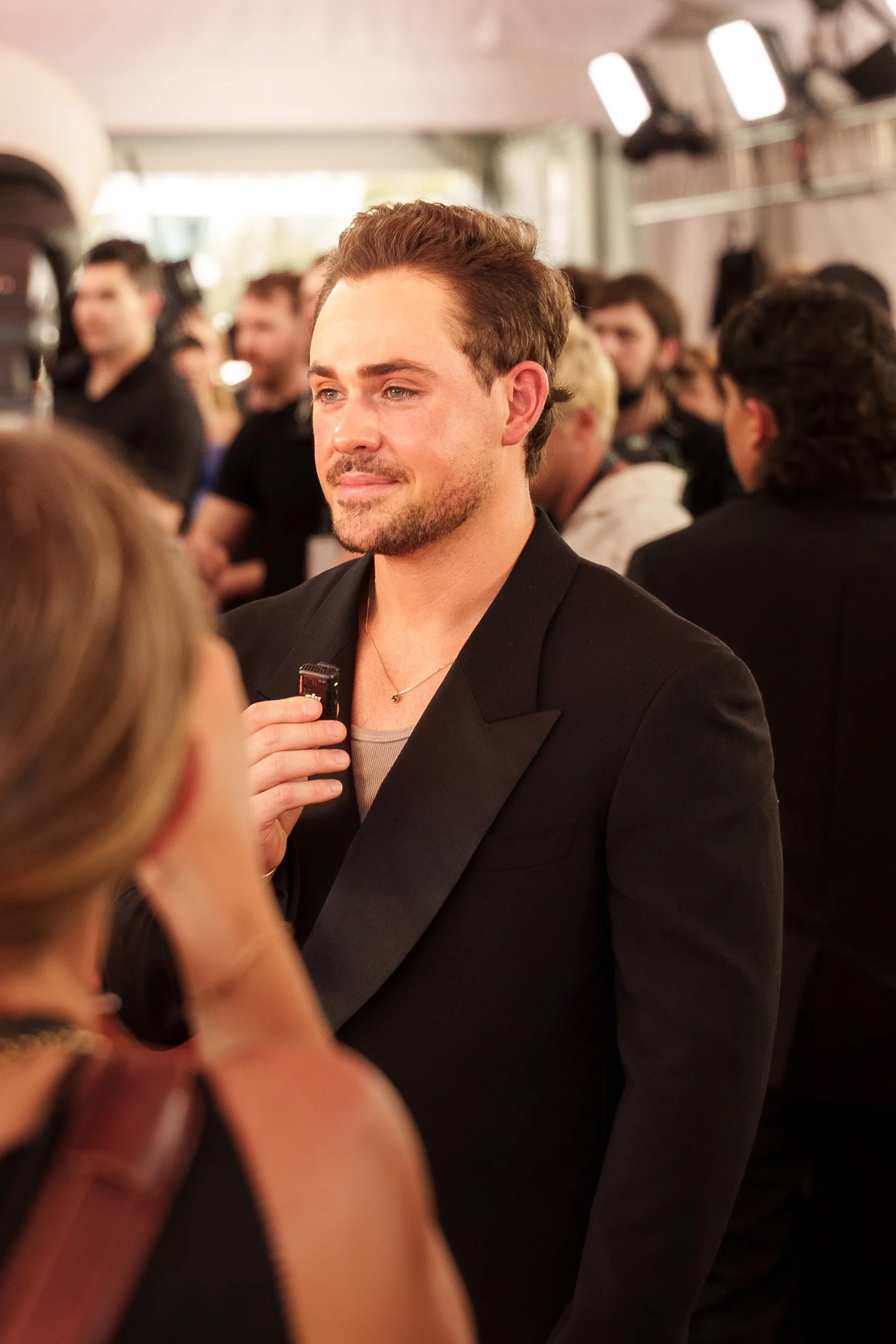 A man with short brown hair and a beard, wearing a black blazer and a gray shirt, is standing against a blurred background of people at an event, holding a small device near his chest with a slight smile.