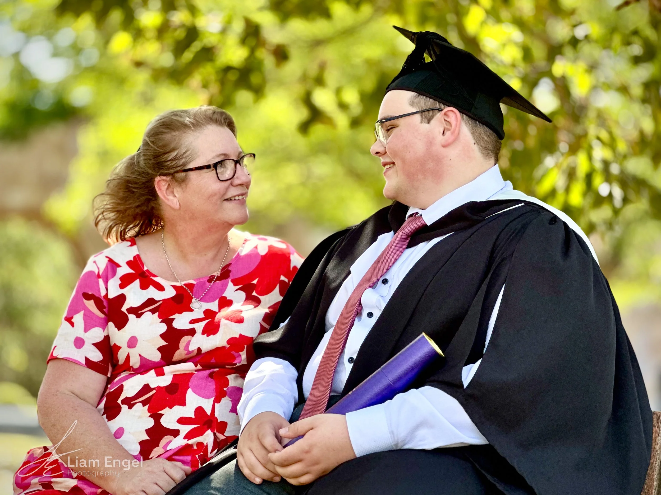 A woman in a red and pink floral dress smiling at a man in a graduation cap and gown, holding a diploma, outdoors with green trees in the background.