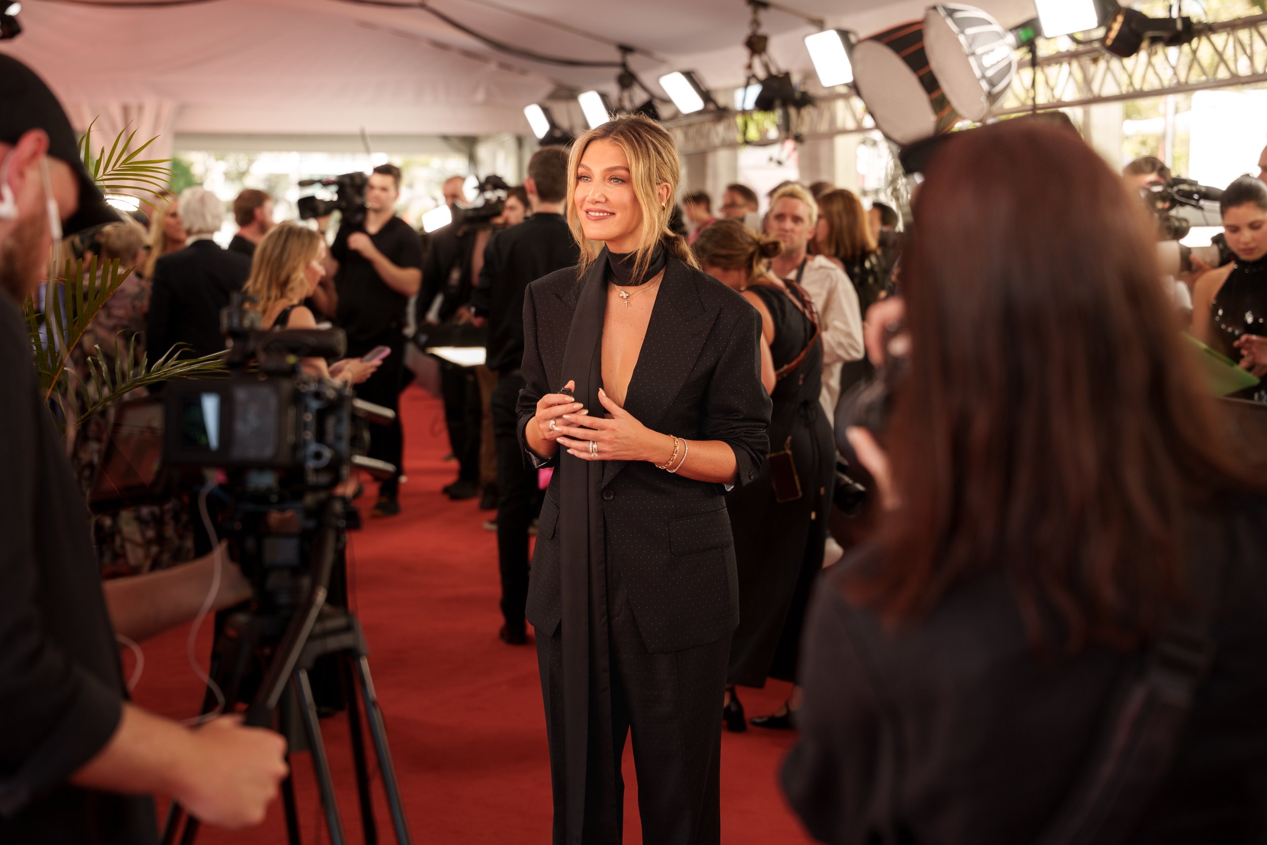 A woman in a black suit with a deep neckline is being interviewed on a red carpet at a formal event, with photographers and other attendees in the background.