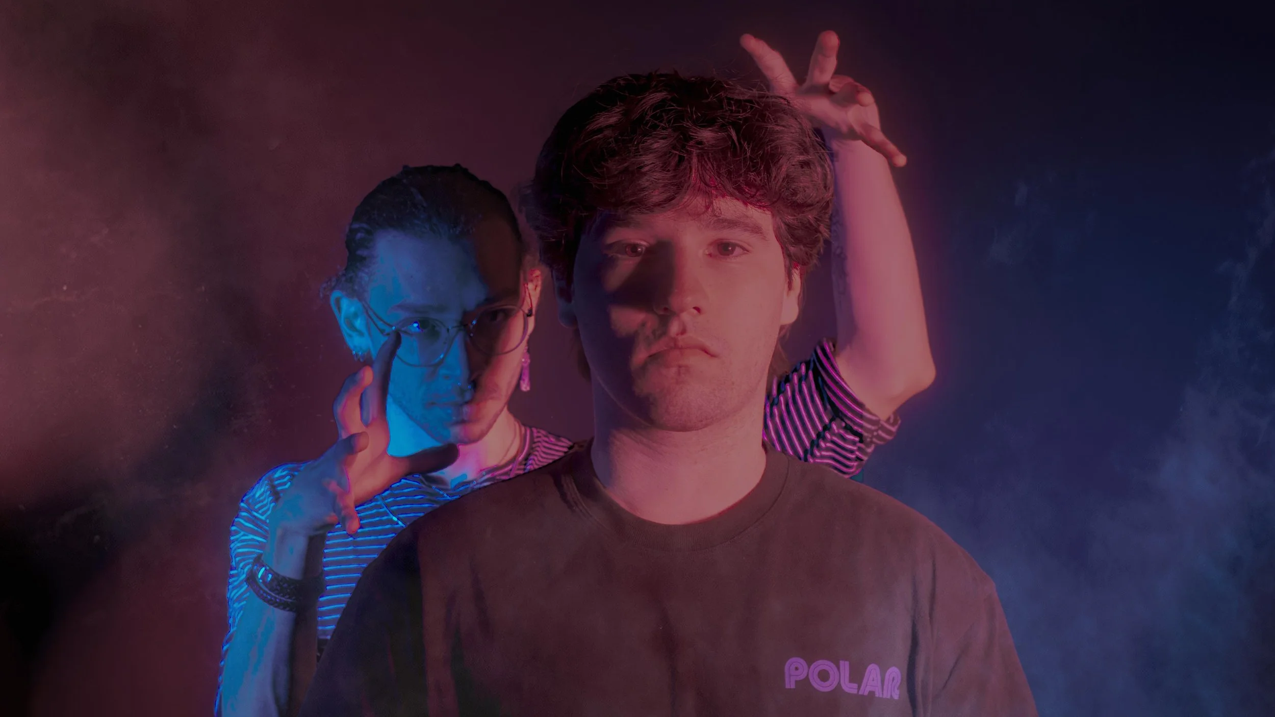 Two young adults, a woman and a man, are standing in a colorful smoky environment. The woman is wearing glasses and earrings, making a gesture with her hand near her face. The man stands in front, looking directly at the camera with a neutral express