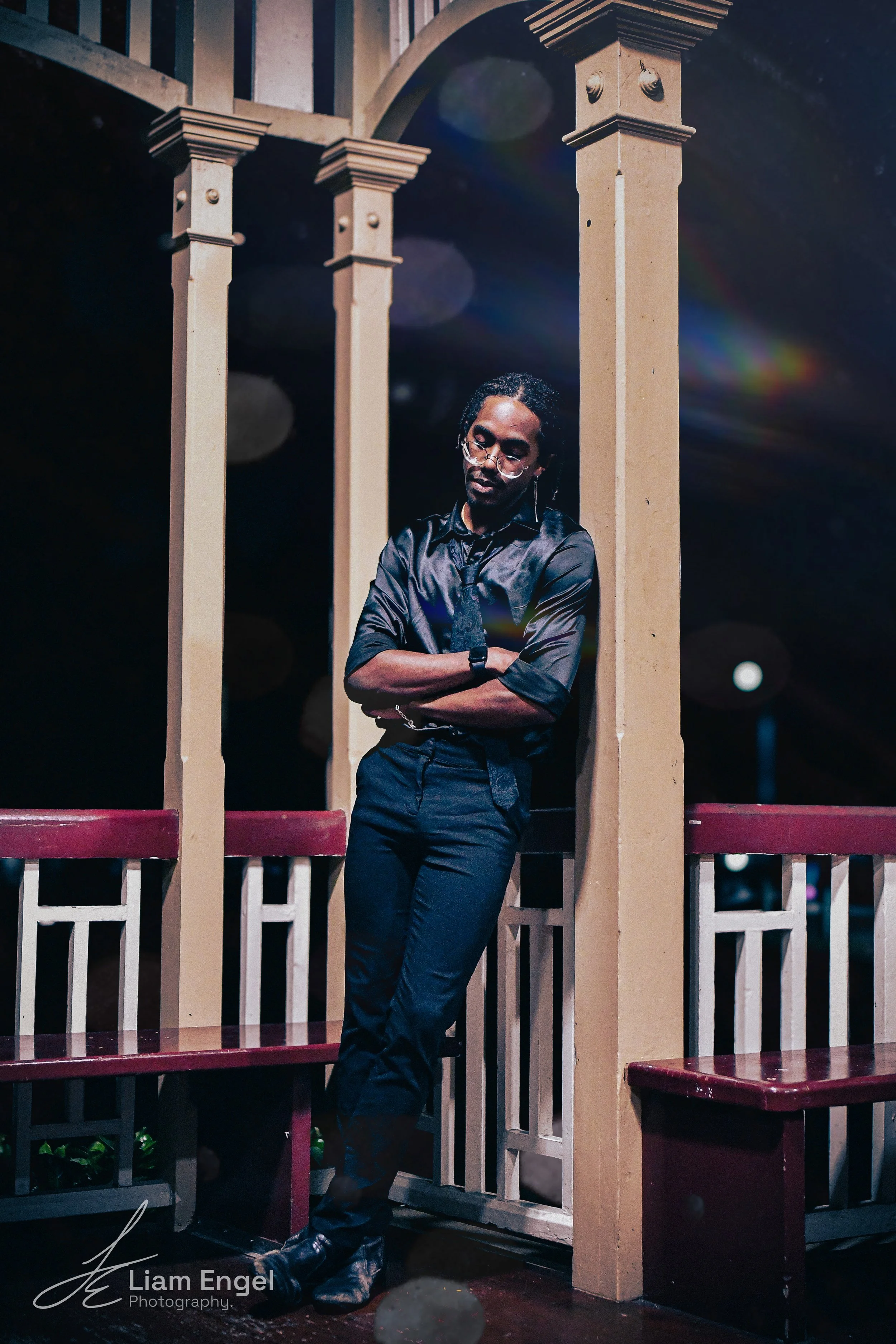 A man with glasses and dreadlocks, dressed in a black shirt and black pants, standing with arms crossed on a wooden structure at night, with dark sky and bokeh lights in the background.
