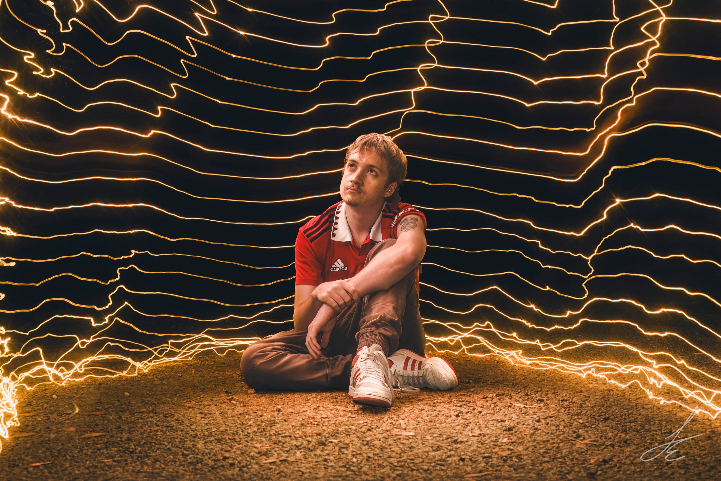 A young man sitting on the ground at night, surrounded by orange and yellow lightning effects created with long exposure photography, smiling and wearing a red Adidas shirt, brown pants, and sneakers.