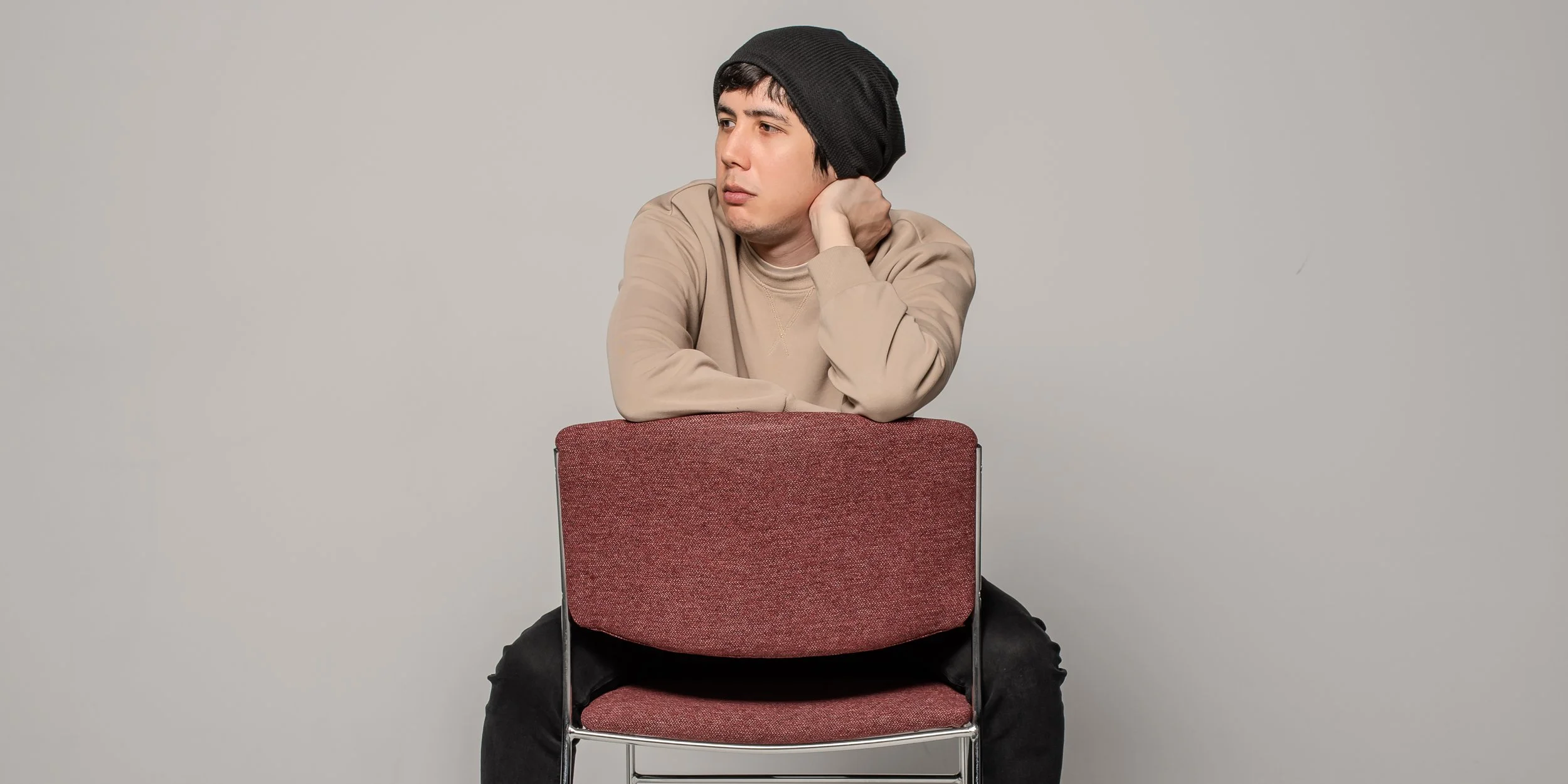 Young man sitting on a red chair with his arms resting on the backrest, wearing a beige sweatshirt and a black beanie, gazing to the side with a thoughtful expression against a plain light gray background.