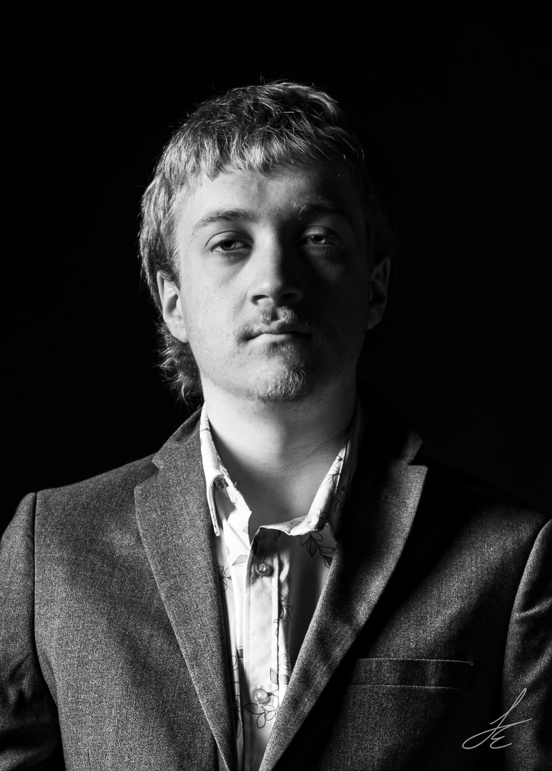 Black and white portrait of a man wearing a suit jacket and a collared shirt with a floral pattern, looking slightly to the side with a serious expression.