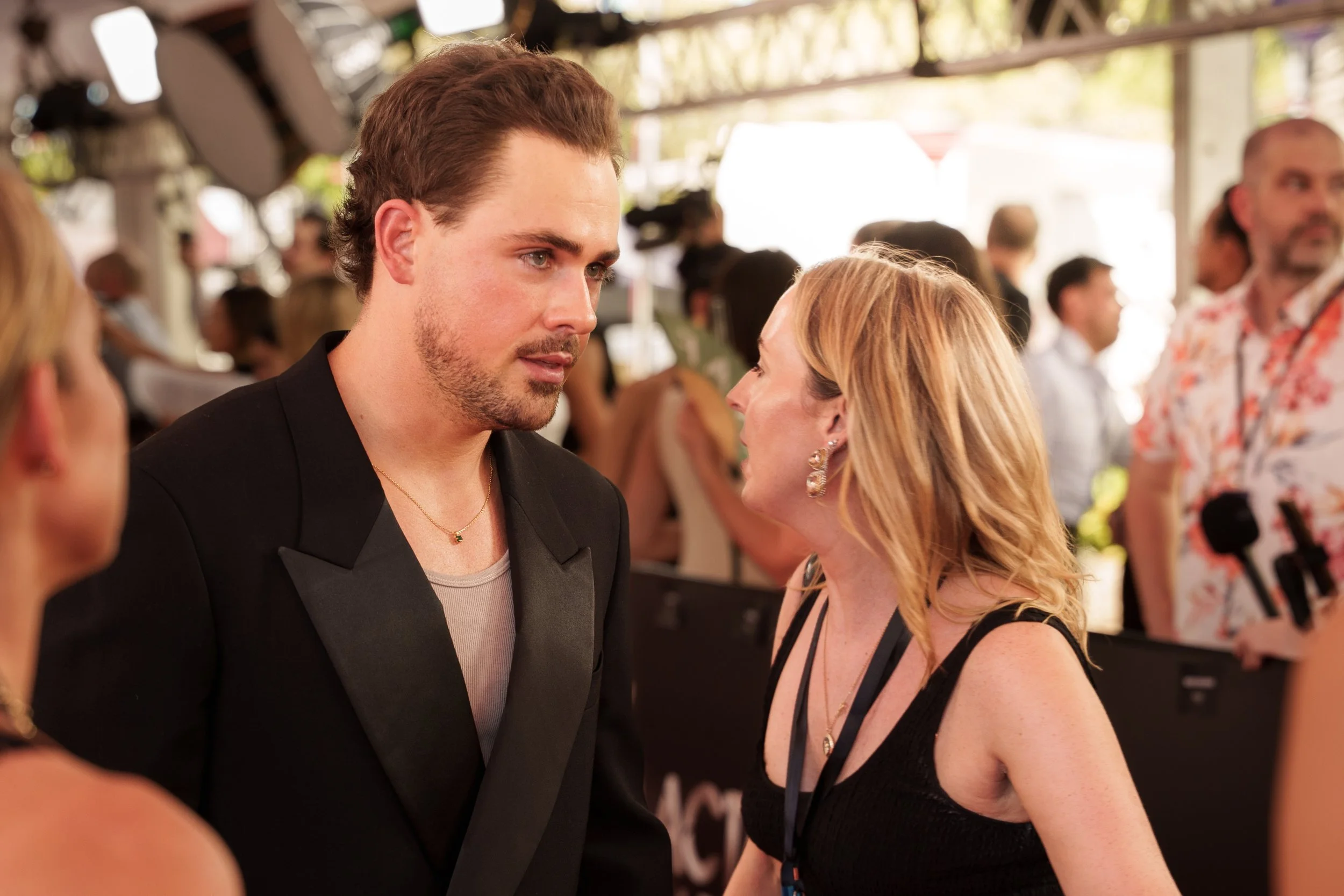 A man and a woman face each other in conversation at a social event, with several other people in the background.