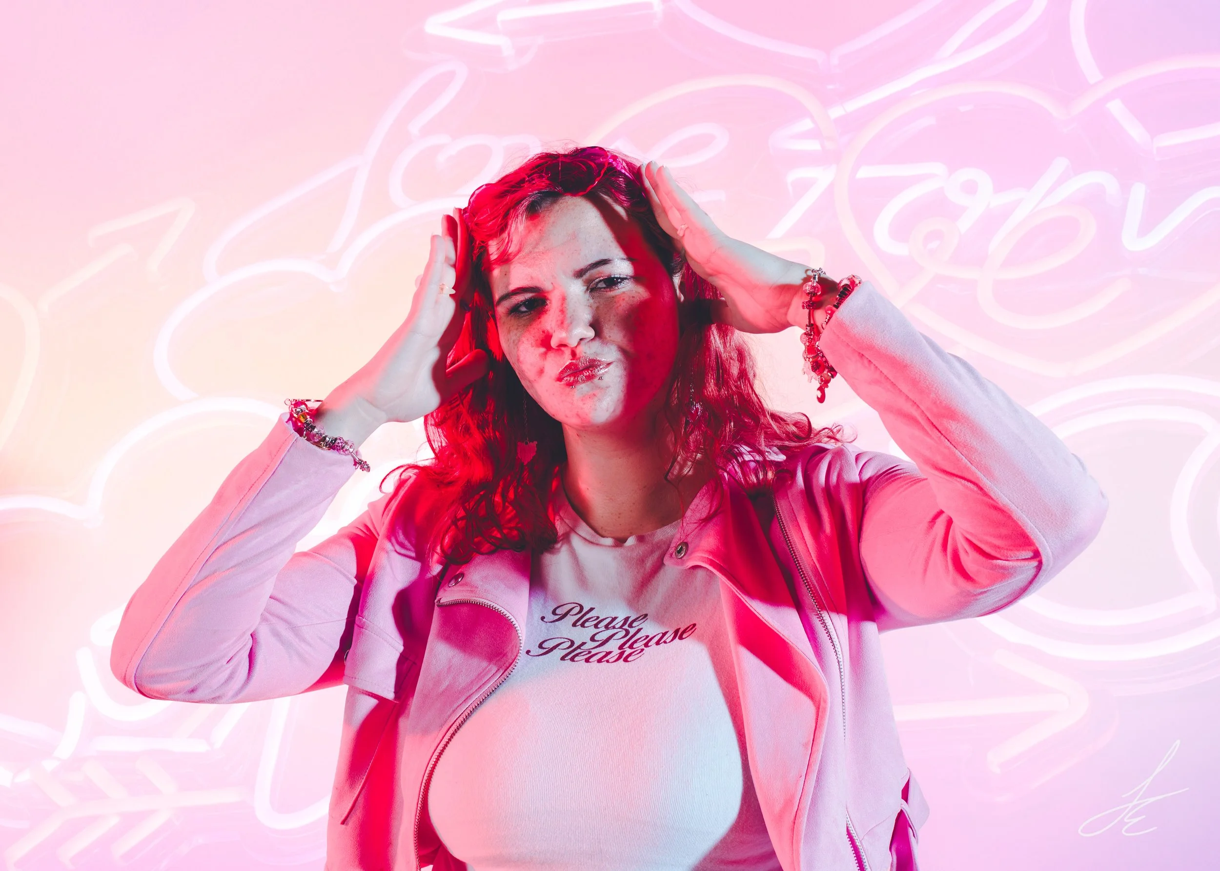 A young woman with red hair, wearing a pink jacket and white shirt with the words 'Please Please Please', making a playful facial expression, touching her head, with pink and purple neon signs in the background.