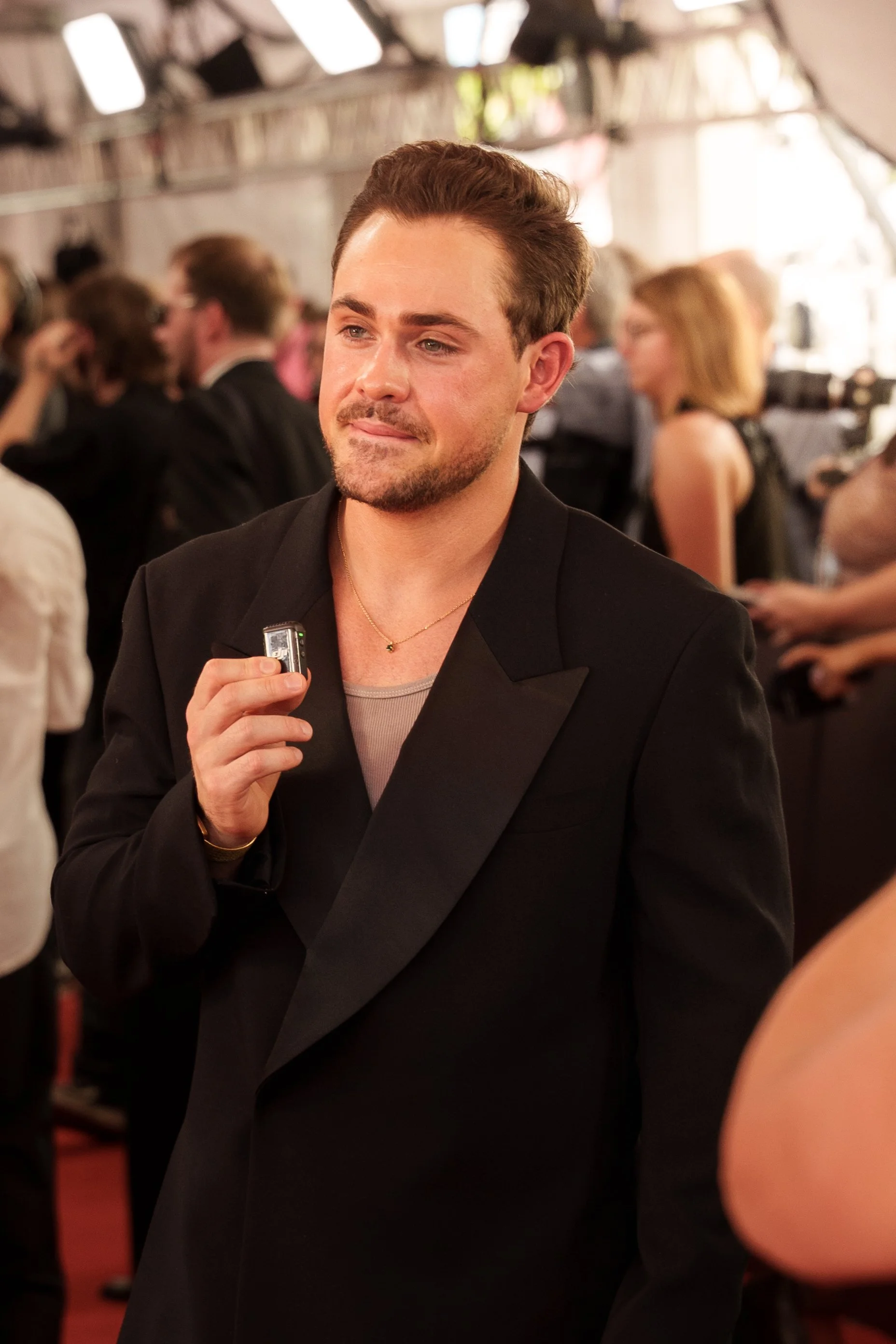 A man in a black tuxedo holding a microphone at a social event, with other people in the background in a well-lit, rustic setting.