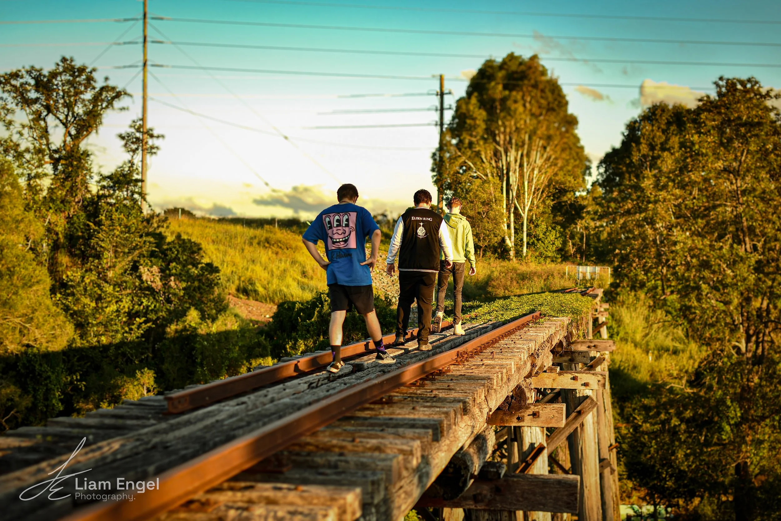 Three young men walking on an old wooden train bridge during sunset, surrounded by green trees and power lines in the background.