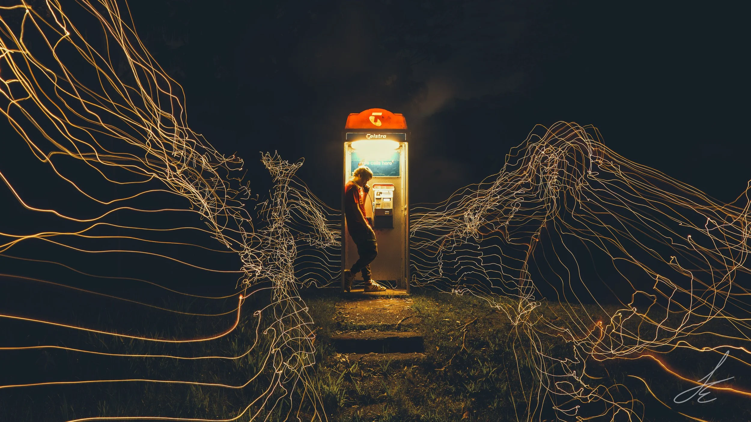 A person standing inside a brightly lit phone booth at night, with light trails surrounding the booth creating a dynamic, swirling pattern against a dark sky.