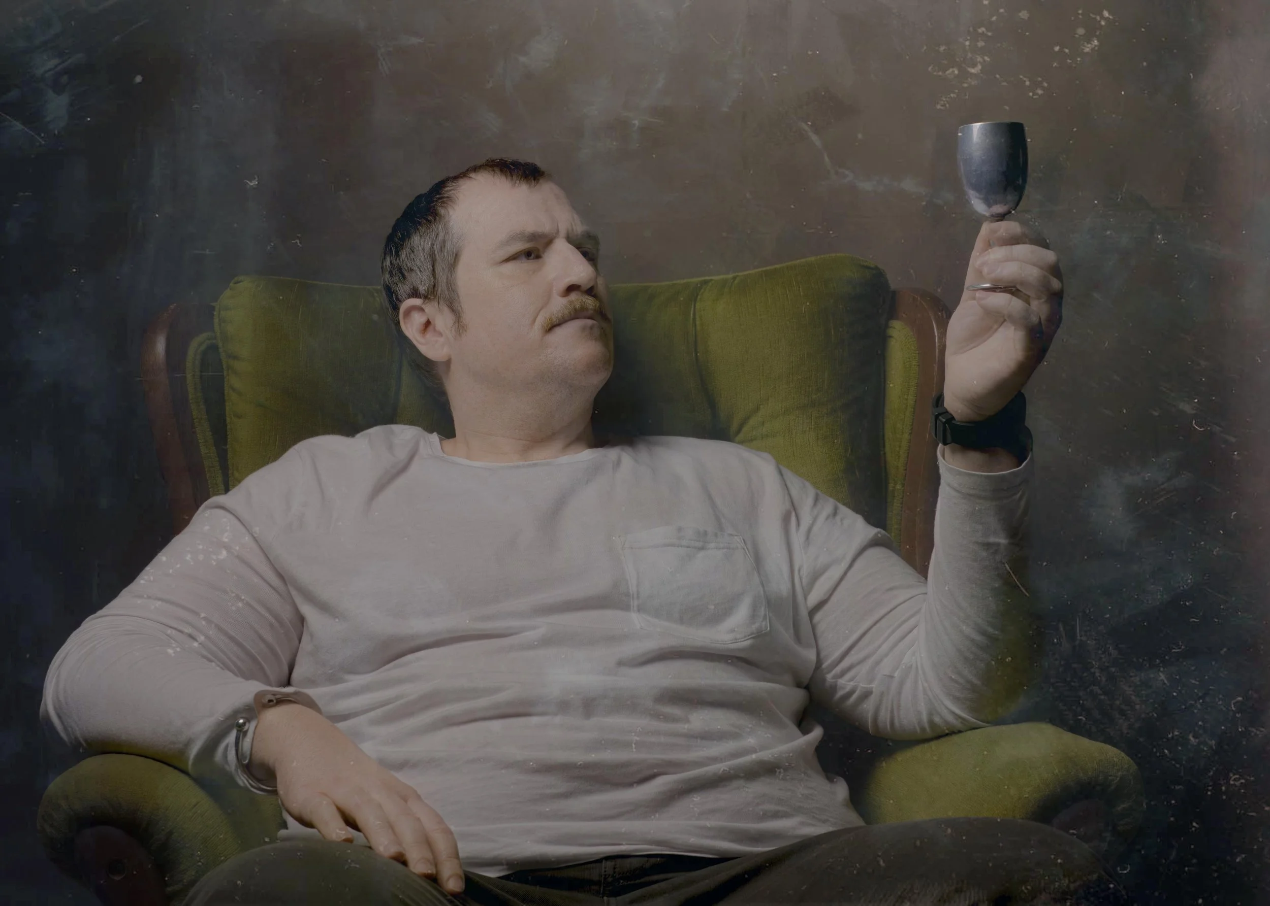 Man lying on a green vintage armchair, holding a wine glass, with a contemplative expression.