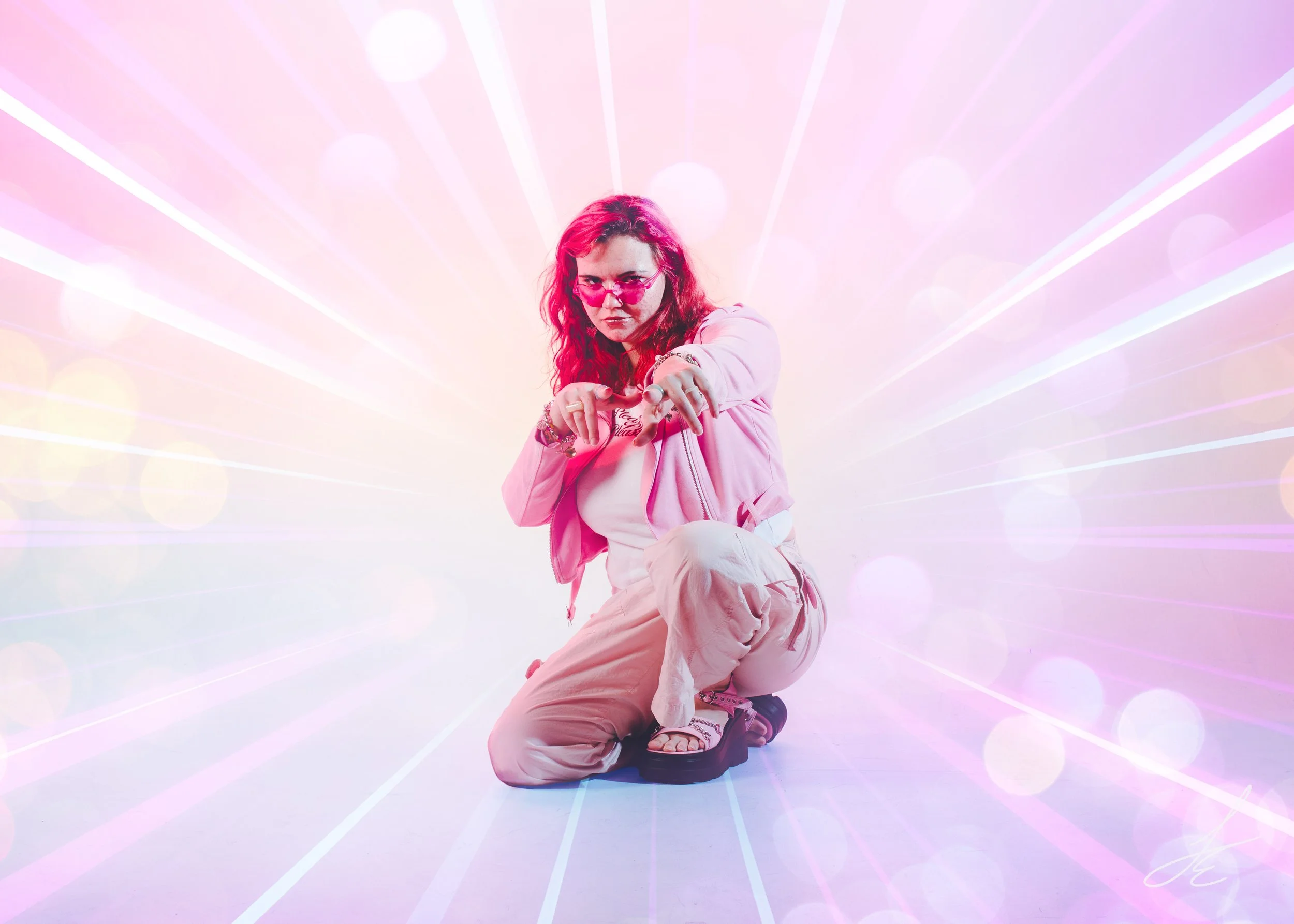 A woman with red hair, pink sunglasses, and pink clothing kneeling on the floor pointing towards the camera with a colorful, futuristic background of pink, purple, and white light rays and bokeh effect.