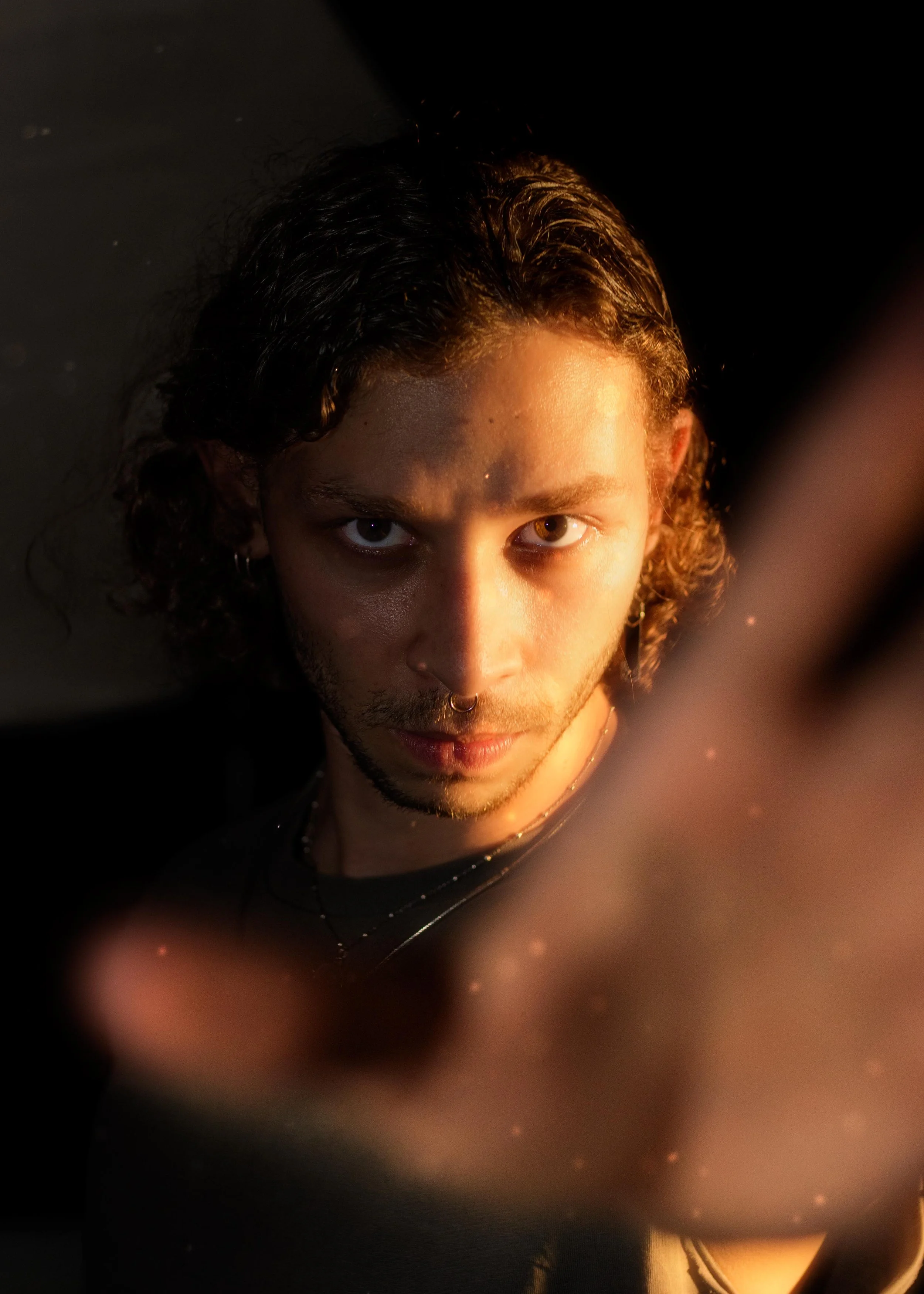 A young man with long, wavy hair, piercing eyes, and facial piercings, reaching towards the camera against a dark background with warm lighting.