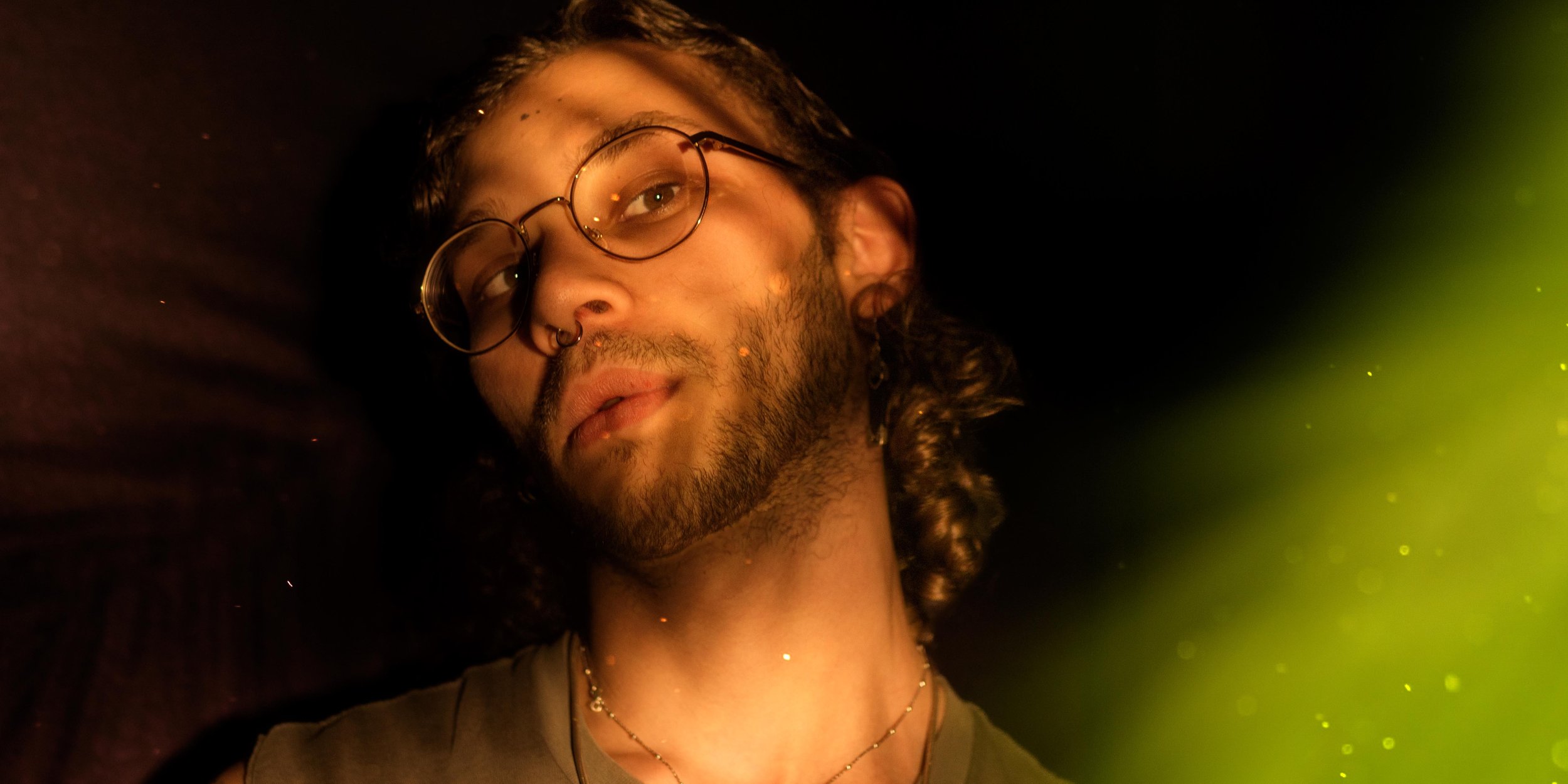 A young man with glasses, facial hair, and long curly hair, lying against a black background with a greenish light on the right side.