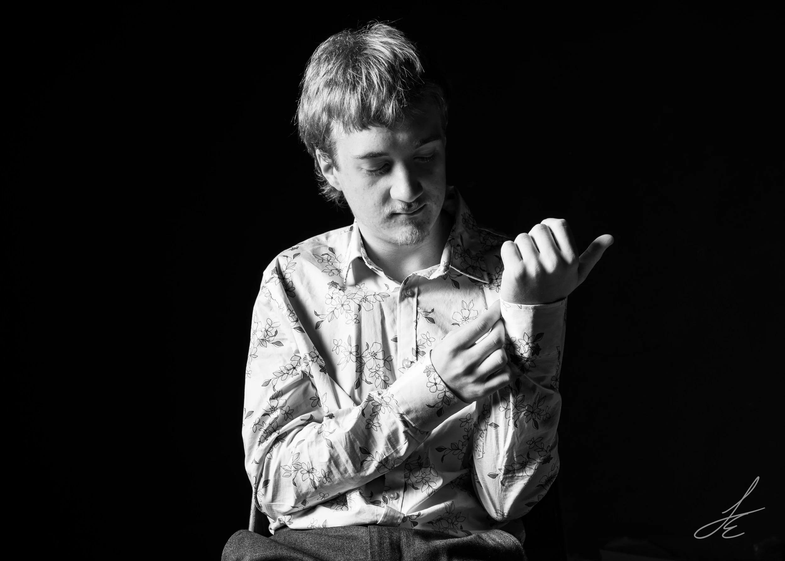 A black-and-white photo of a young man with light hair and a light beard, wearing a floral-patterned shirt, looking down with a thoughtful expression. His arms are crossed, with one hand adjusting his sleeve, set against a dark background.