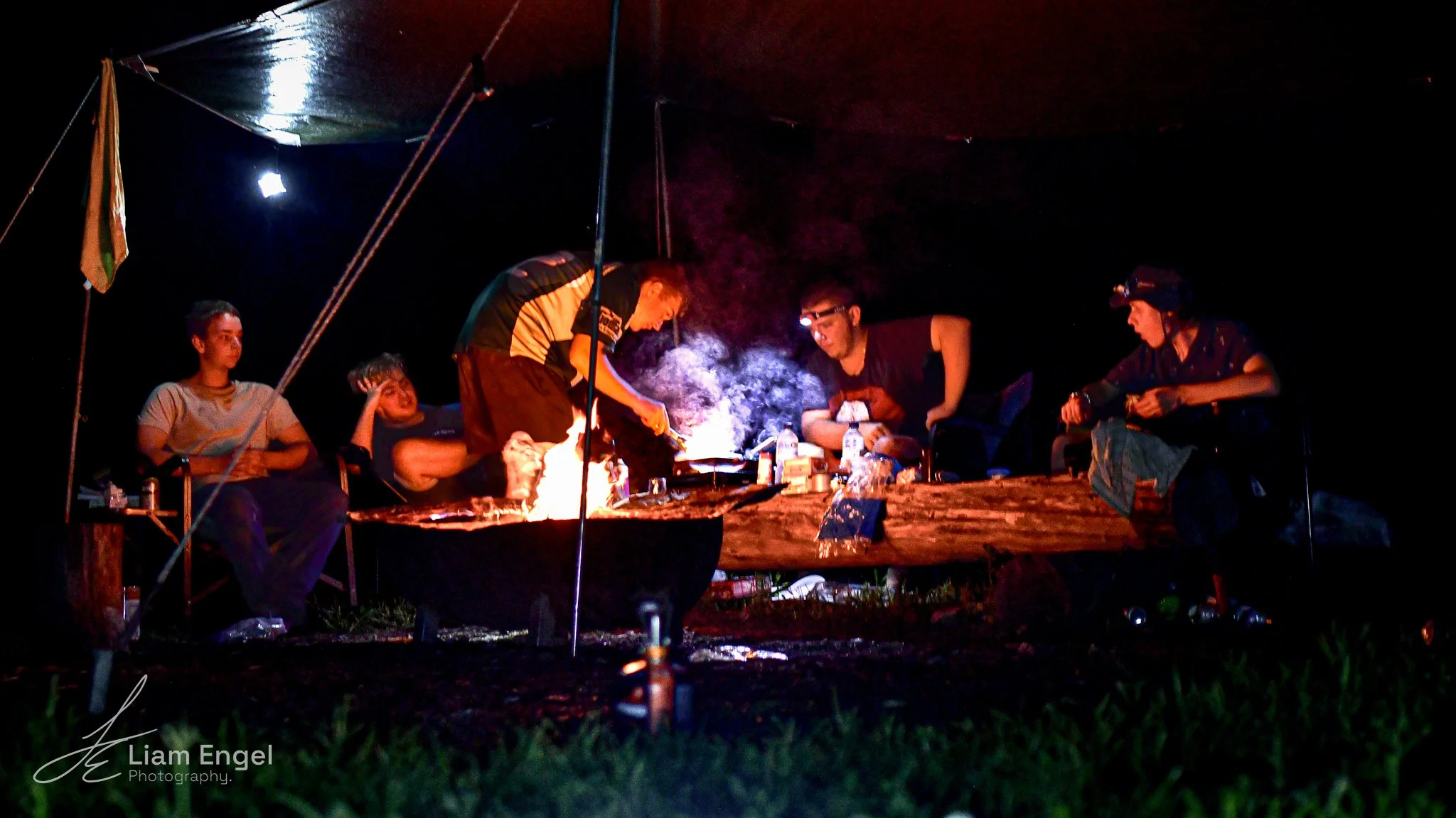 Group of five people camping around a fire in a tent at night, with two cooking over a campfire and three sitting nearby, some with headlamps on.