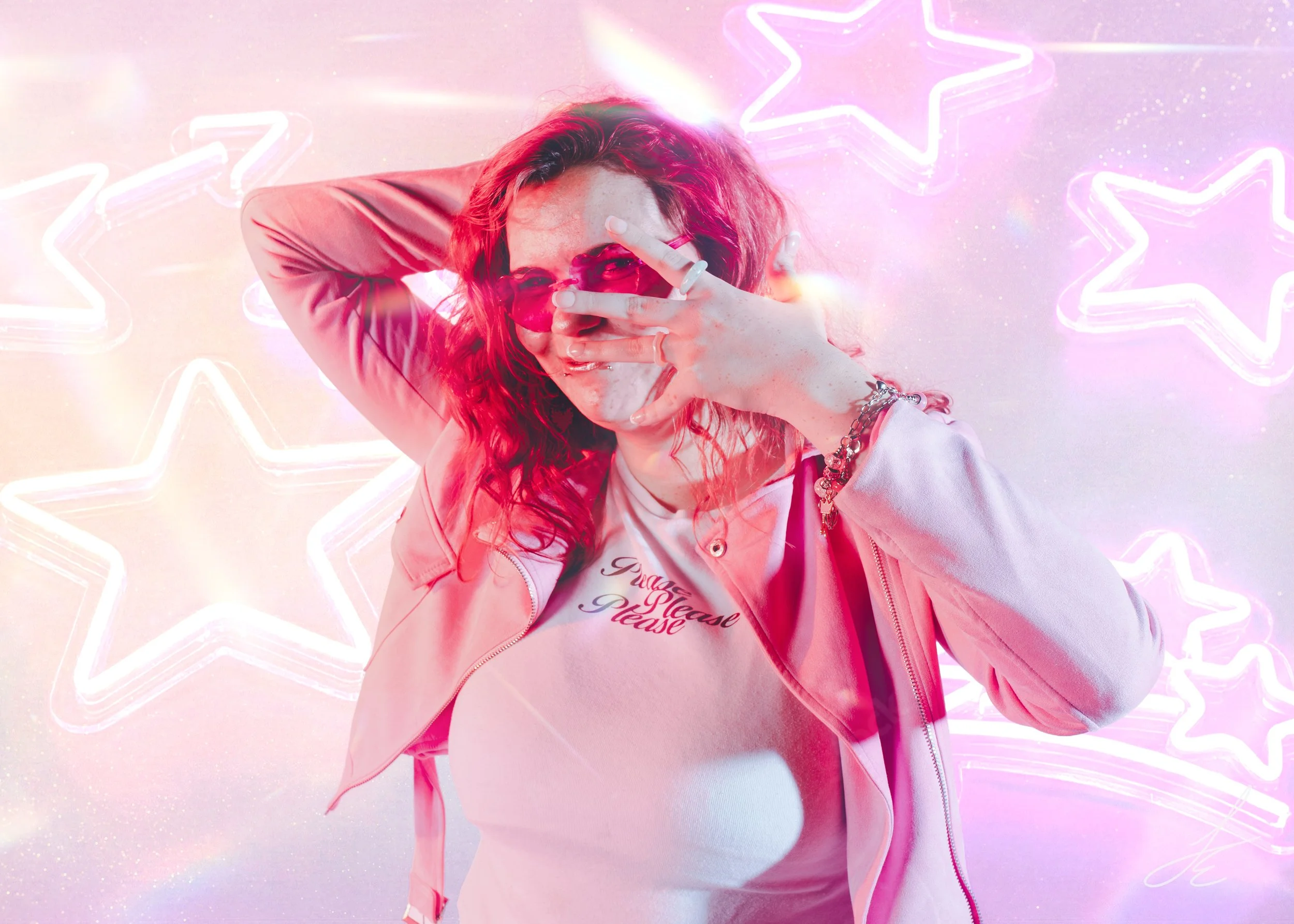 A woman with curly hair posing in front of a neon pink and purple star-shaped background, wearing pink sunglasses, a pink jacket, and a white t-shirt with red writing, covering part of her face with her hand.