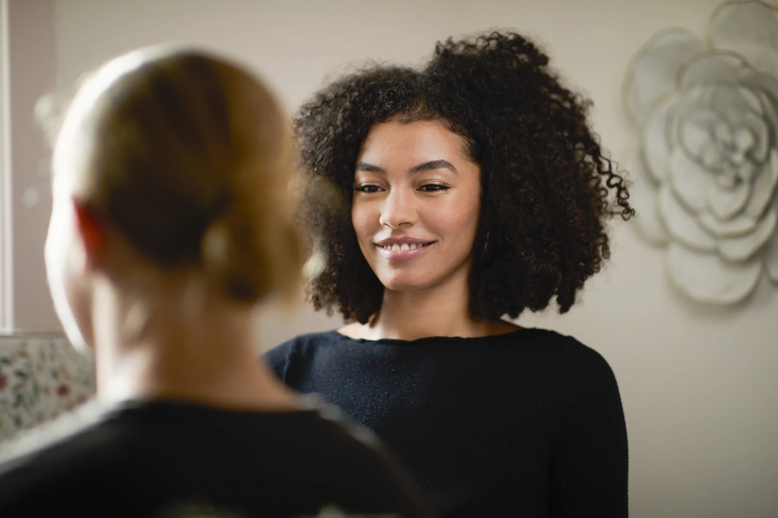 A woman with curly hair smiling and looking at another person who has a bun hairstyle, in a well-lit indoor setting with wall art in the background.