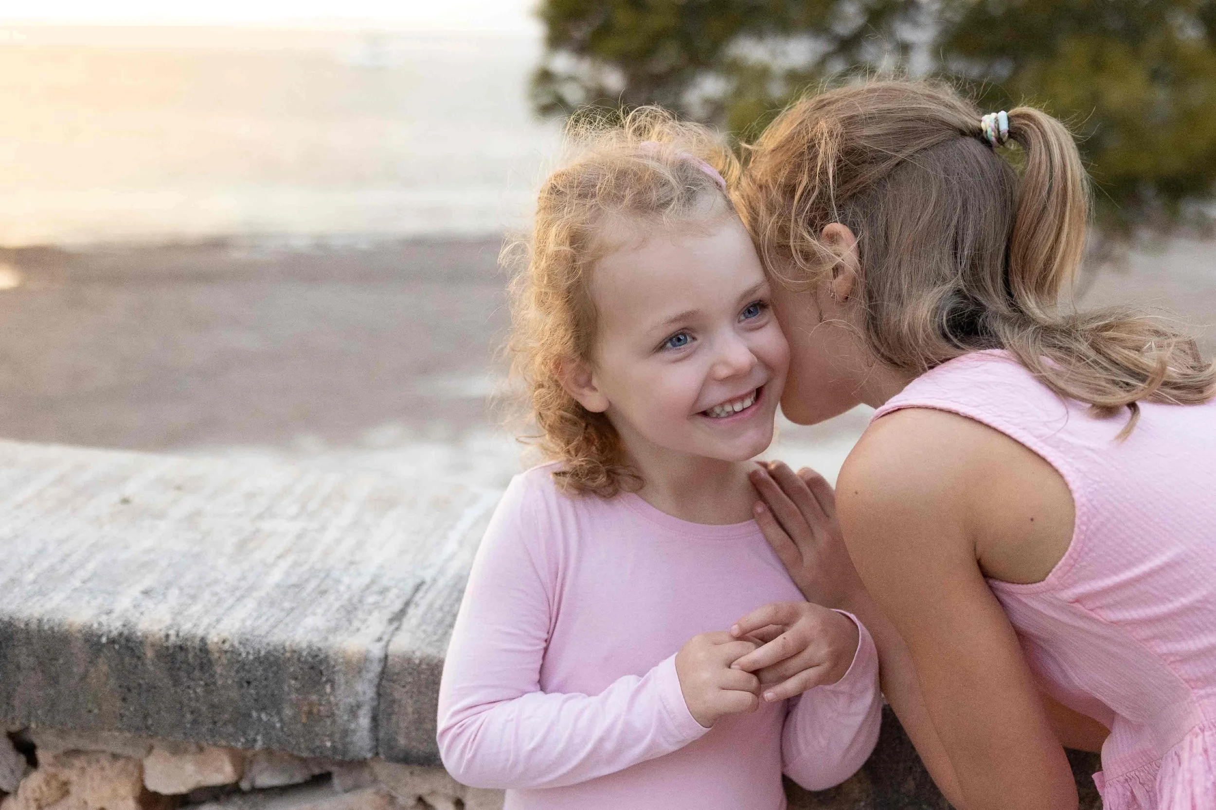 Familien- & Kinderfotografie in Hamburg und Umgebung von Klaudia Schmidt. Zwei Mädchen in pinken Kleidern, eines flüstert dem anderen etwas ins Ohr bei Sonnenuntergang im Freien.