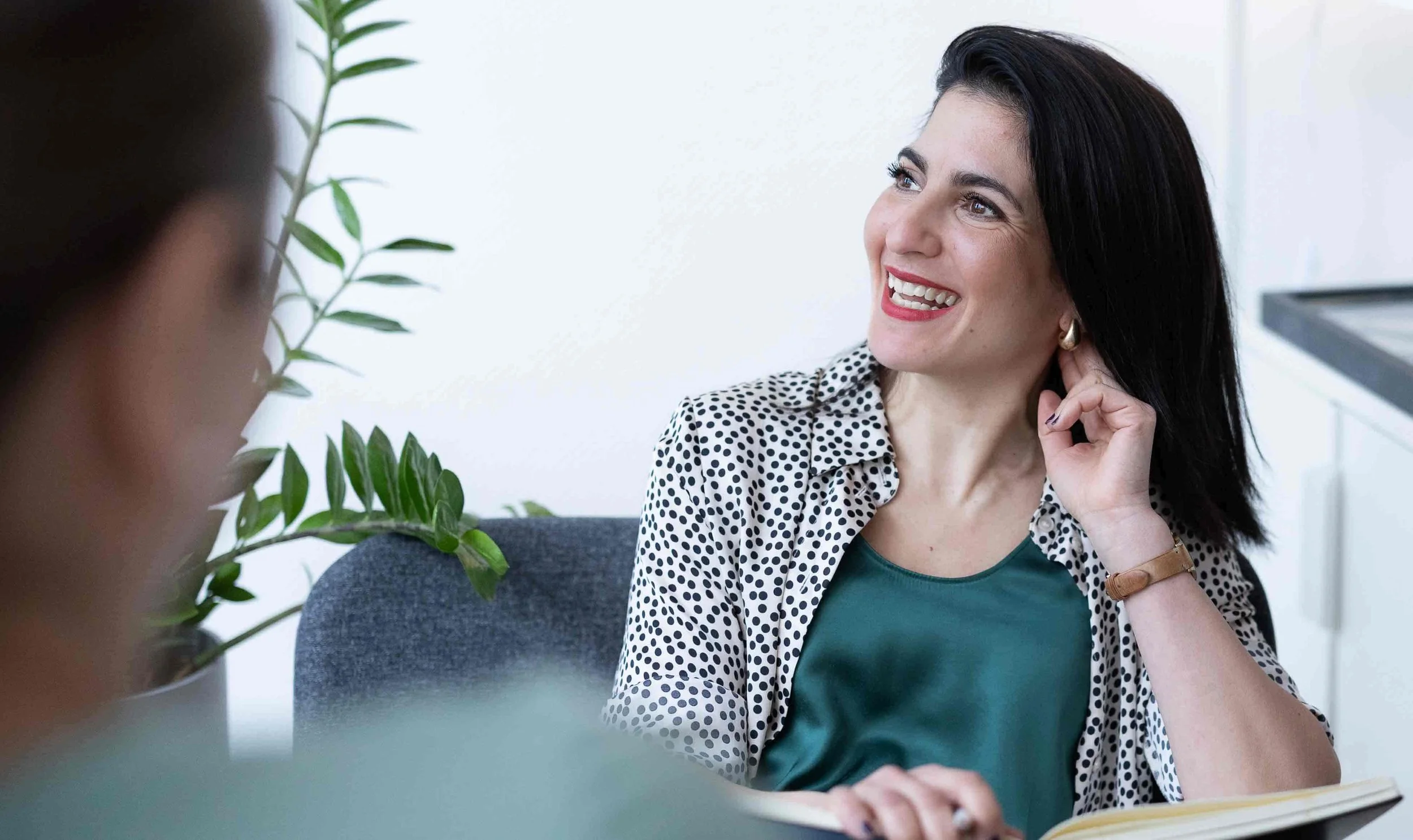 Professionelle Businessfotografie in Hamburg und Umgebung von Klaudia Schmidt. Eine lächelnde Frau im Gespräch, sitzt an einem Schreibtisch, neben ihr eine Pflanze, im Hintergrund ist eine weiße Wand.