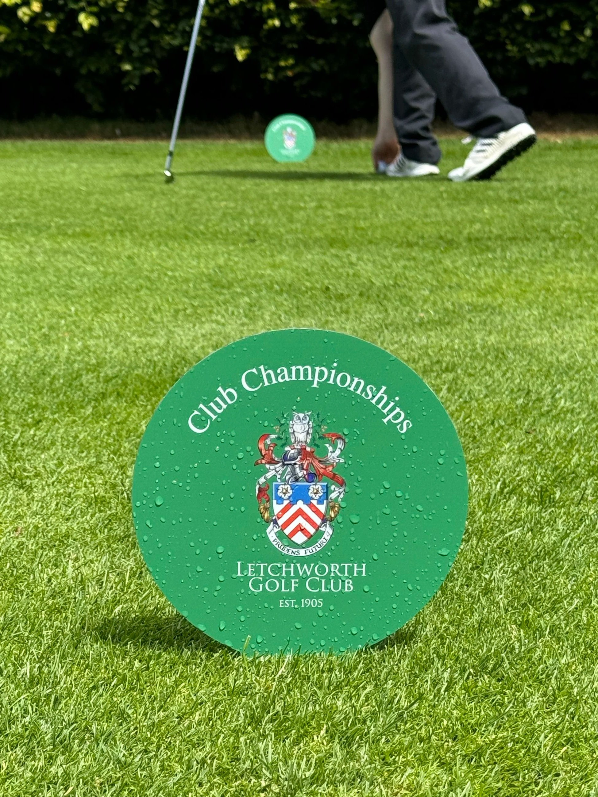 Green medallion reading 'Club Championships Letchworth Golf Club EST 1905' with a crest, placed on a golf course green, with a golfer in the background preparing for a shot.