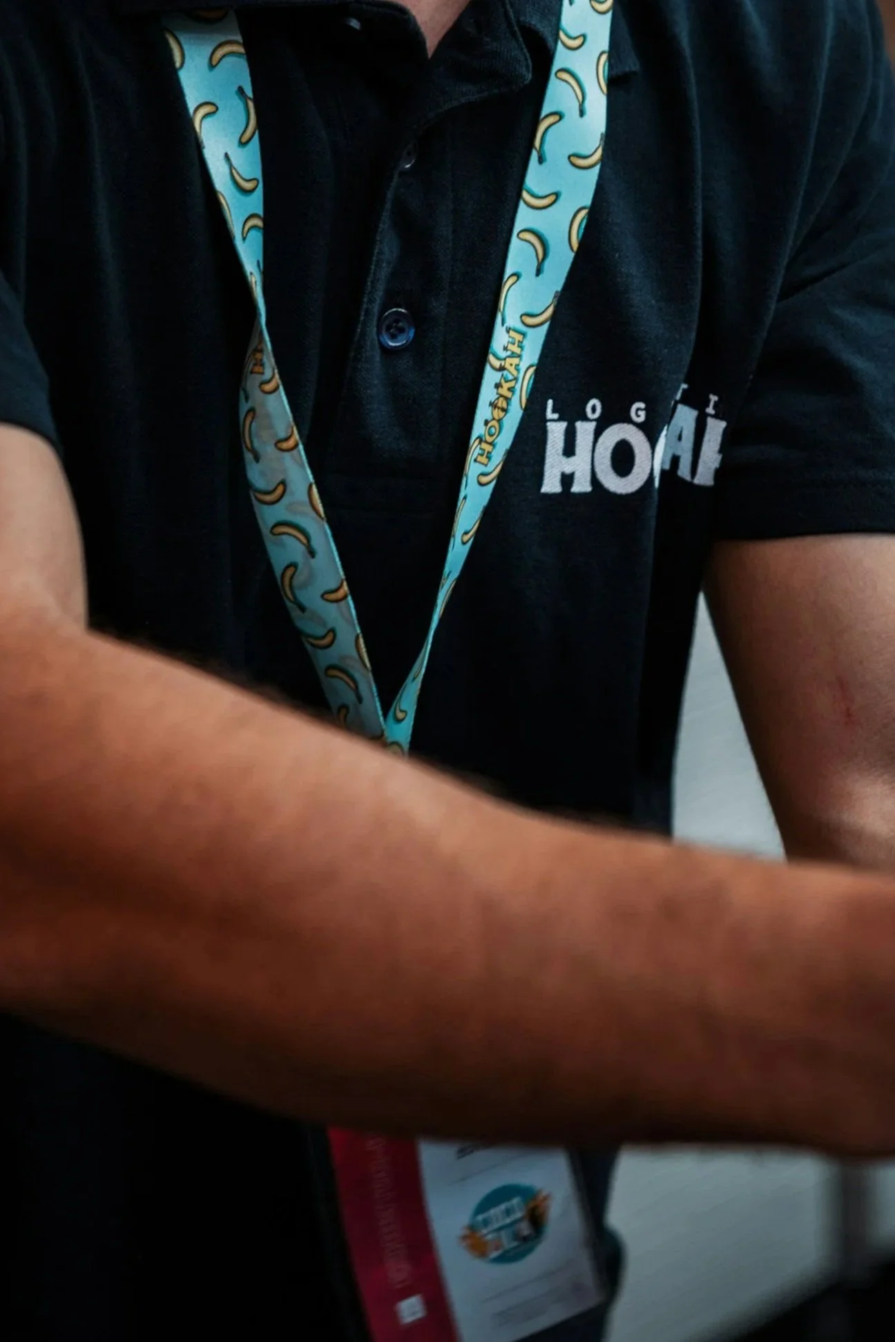 Close-up of a person wearing a black polo shirt with a blue camouflage lanyard and apron featuring a banana pattern. The shirt has the words 'LOG HOAI' printed on the chest.