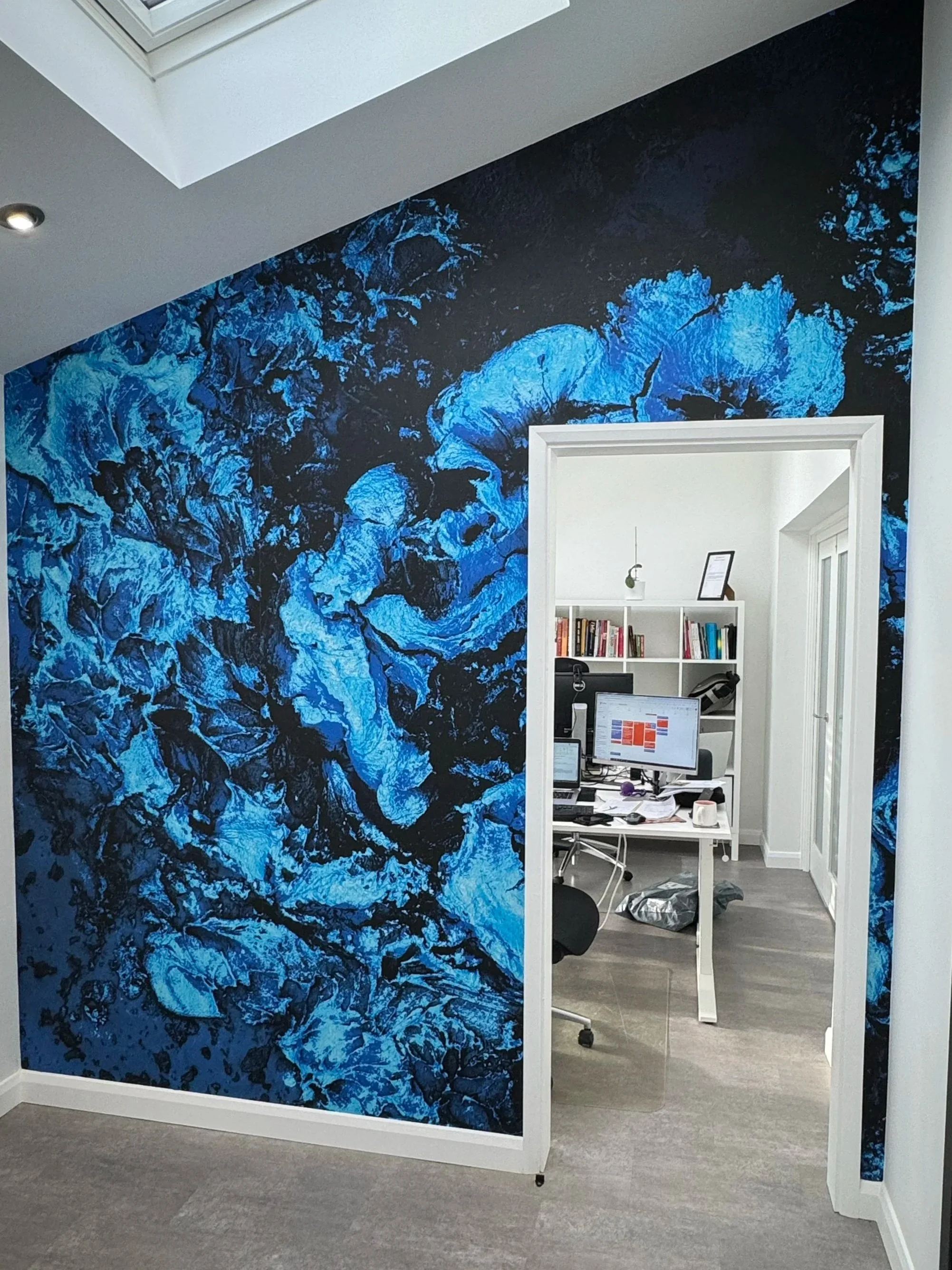 Office room with a wall featuring a blue and black abstract mural, and a doorway leading to a workspace with a desk, computer, bookshelves, and various office supplies.