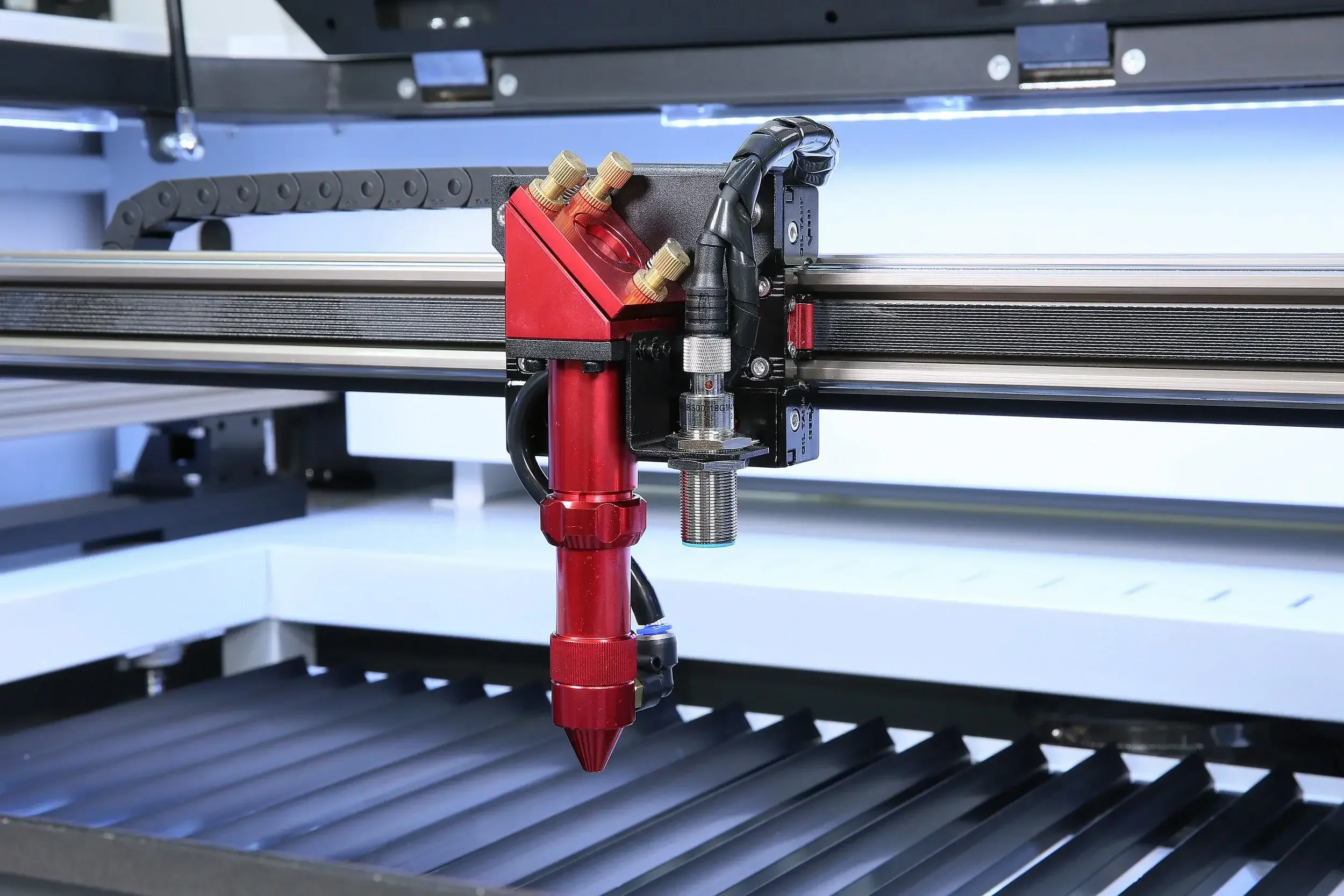 Close-up view of a red industrial nozzle attached to a metal structure, with hoses connected, likely used for manufacturing or 3D printing.