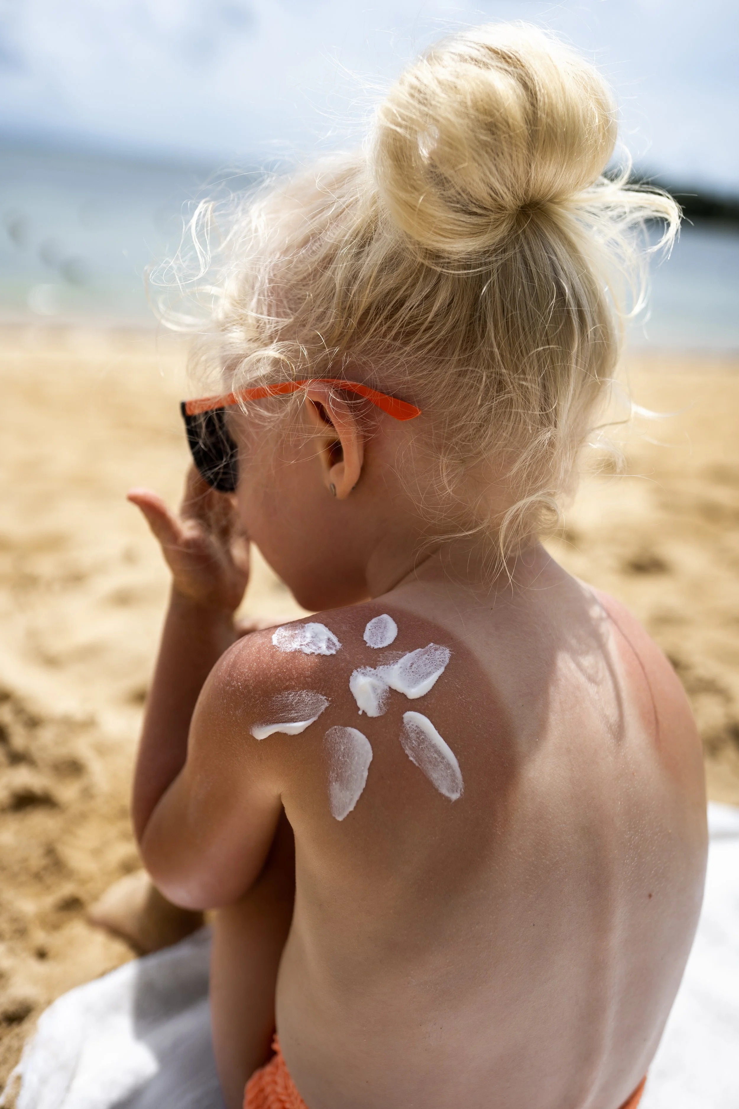 Junge Frau mit Sonnenbrille sitzt am Strand, Sonne auf der Haut, Blick zum Meer.