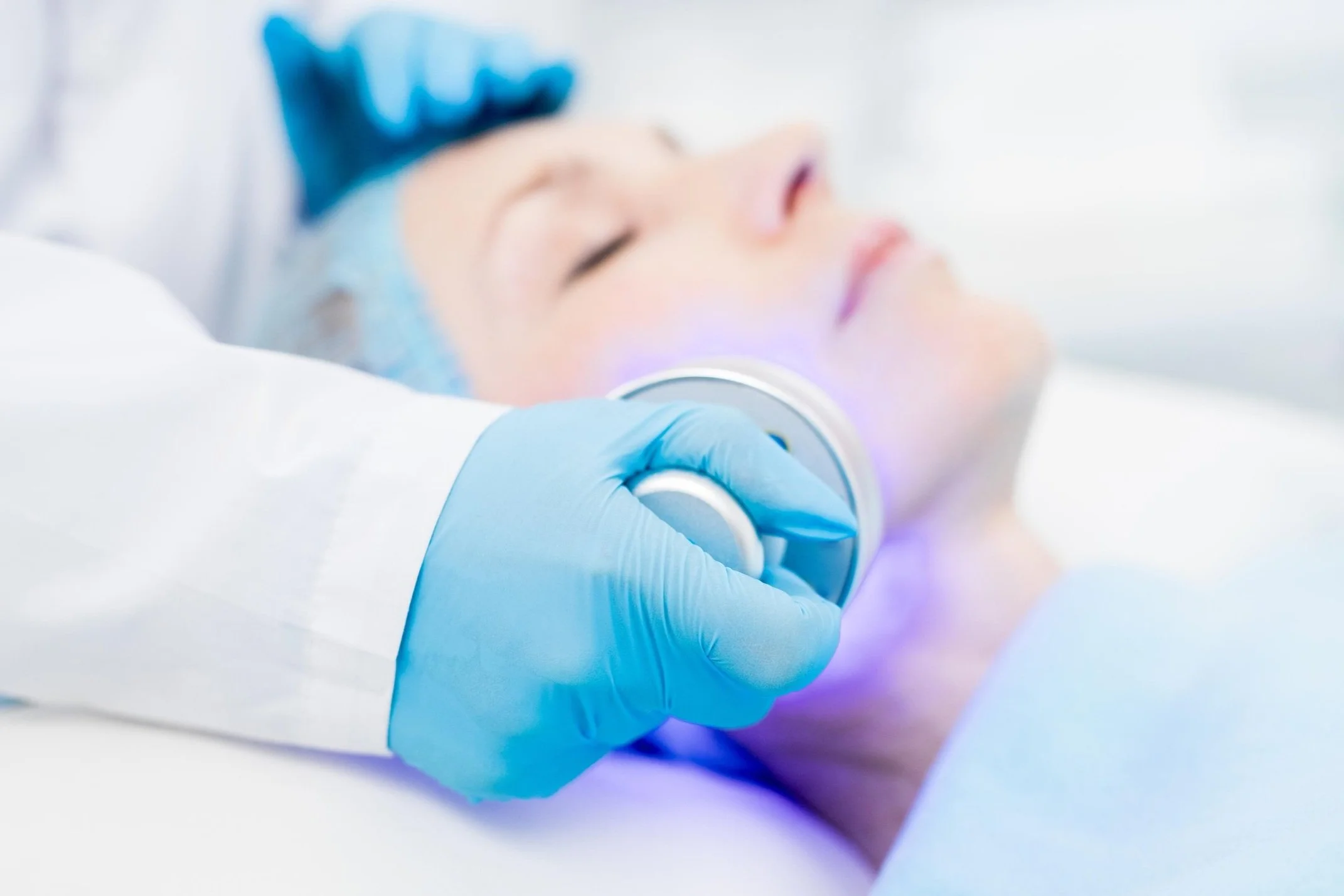 Ärztin benutzt ein Gerät mit blauer LED-Licht auf das Gesicht eines liegenden Patienten mit geschlossenen Augen, in einem medizinischen Umfeld.