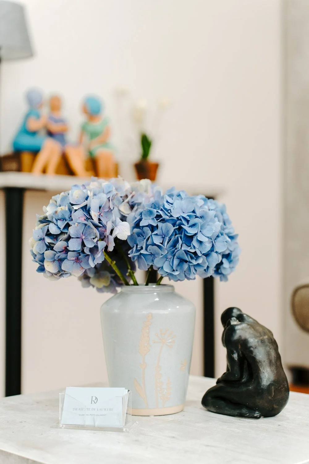 Blue hydrangeas in a decorative vase next to a small black sculpture on a table with a business card holder.