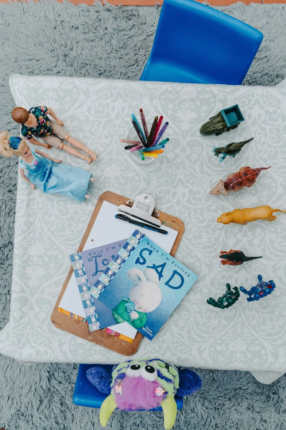 Dr Riette de Lauwere, Clinical Psychologist Childs therapy table which is a top view of a child's play table with Barbie dolls, colorful markers in a cup, toy animals, and children's books titled 'When I'm Feeling Sad' and 'When I'm Feeling Lonely.'