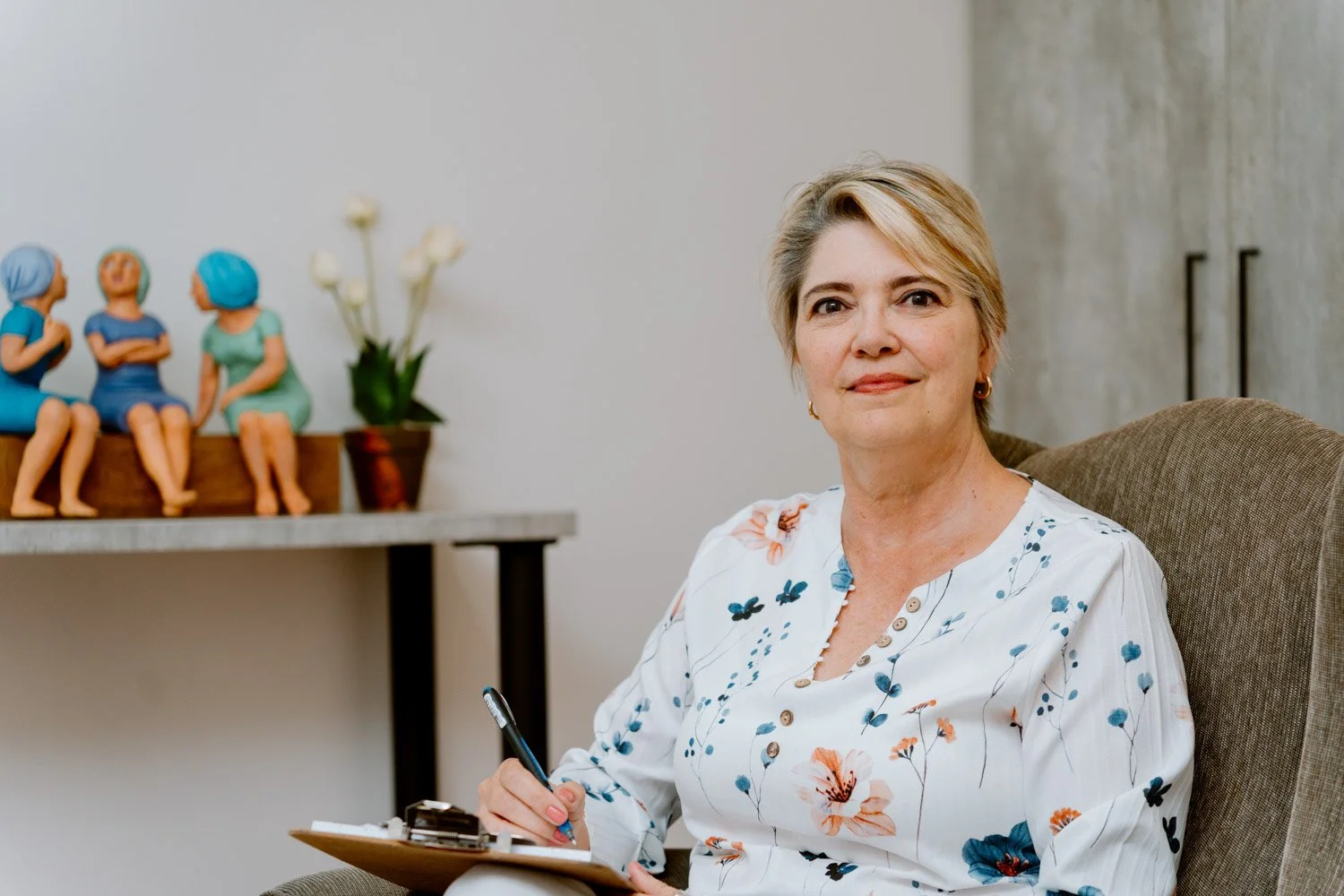 Dr Riette de Lauwere, Clinical Psychologist sitting with a clipboard in an office, wearing a floral blouse, and smiling. There are decorative figures and white flowers on a shelf behind her.