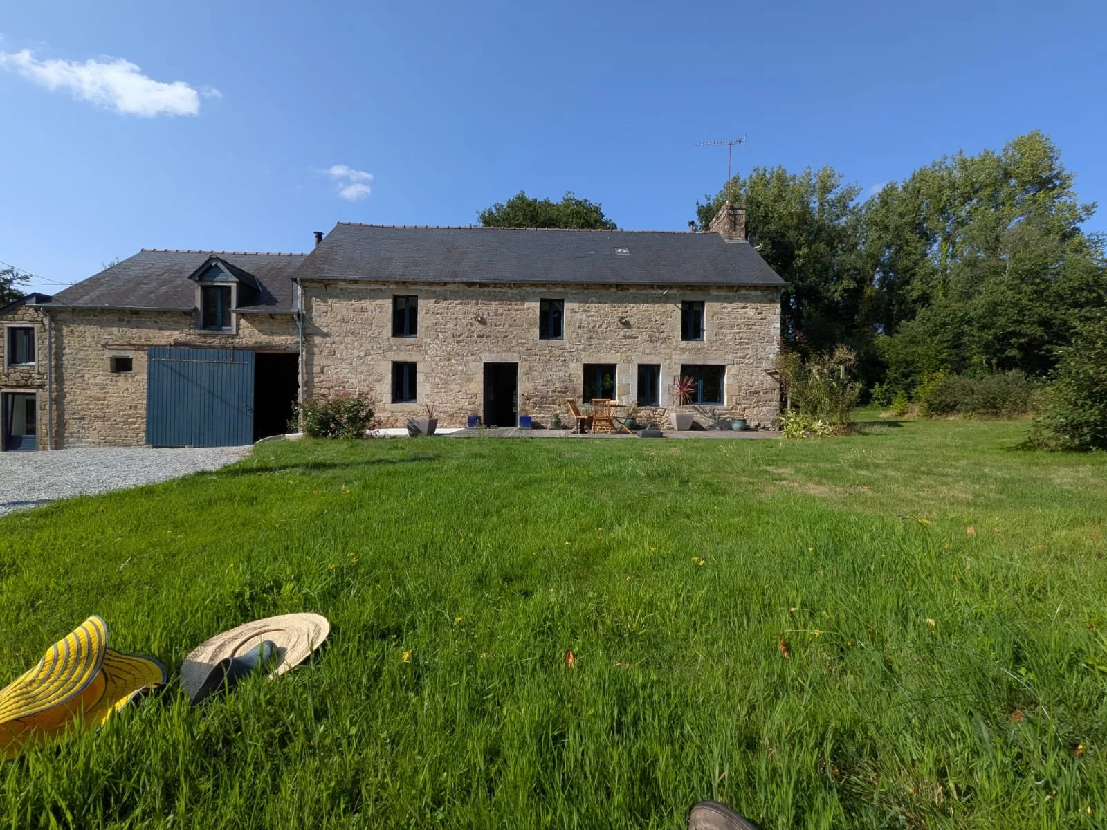 A stone house with a dark roof, multiple windows, and outdoor patio furniture, surrounded by a lush green lawn and trees under a clear blue sky.
