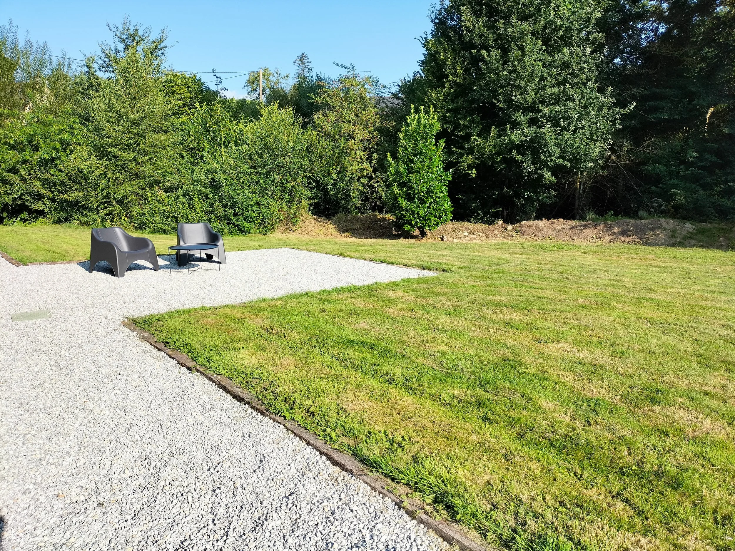 Two black outdoor chairs and a small black table on a gravel patio, with a well-maintained lawn and dense green trees in the background.